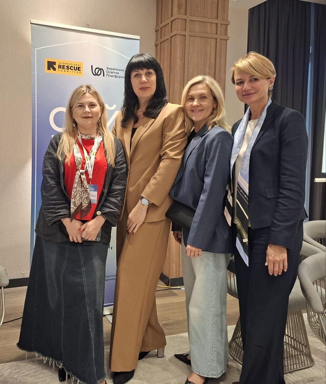 Чотири жінки-учасниці панельної дискусії «Різні громади — спільна мета» в межах проєкту «Обійми 3.0» позують для спільного фото у конференц-залі на тлі інформаційного банера. Присутні вишикувалися в один ряд, посміхаючись у кадр. Друга ліворуч у бежевому брючному костюмі стоїть начальниця служби у справах дітей Херсонської ОДА Тетяна Лушнікова. Поруч із нею розмістилися інші спікерки та представниці БО «Українська освітня платформа»: крайня ліворуч — дівчина у довгій джинсовій спідниці та чорному кардигані з червоним топом, друга праворуч — жінка у темно-синьому піджаку та світлих джинсах, а крайня праворуч — жінка у чорному класичному костюмі зі світлою шийною хусткою. На задньому плані ліворуч встановлено офіційний блакитний банер із логотипами організації «International Rescue Committee» та БО «Українська освітня платформа».
