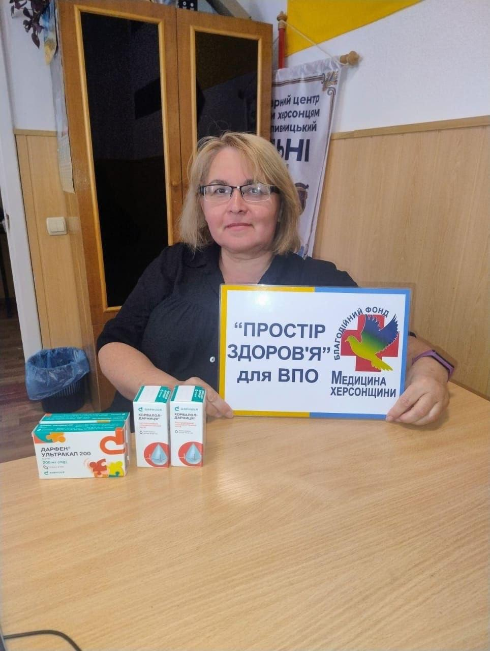 Жінка в окулярах та чорній кофті сидить за столом у кропивницькому координаційному центрі «Вільні разом» та презентує медичну допомогу для ВПО з Херсонщини від БФ «Медицина Херсонщини». Вона тримає в руках табличку з написом «"ПРОСТІР ЗДОРОВ'Я" для ВПО Медицина Херсонщини» та логотипом у вигляді різнокольорового птаха. На дерев'яному столі перед нею розставлені лікарські засоби для переселенців: коробка «Дарфен® Ультракап 200» та два пакування крапель «Корвалол-Дарниця». На задньому плані над плечем жінки висить офіційний банер гуманітарного хабу.