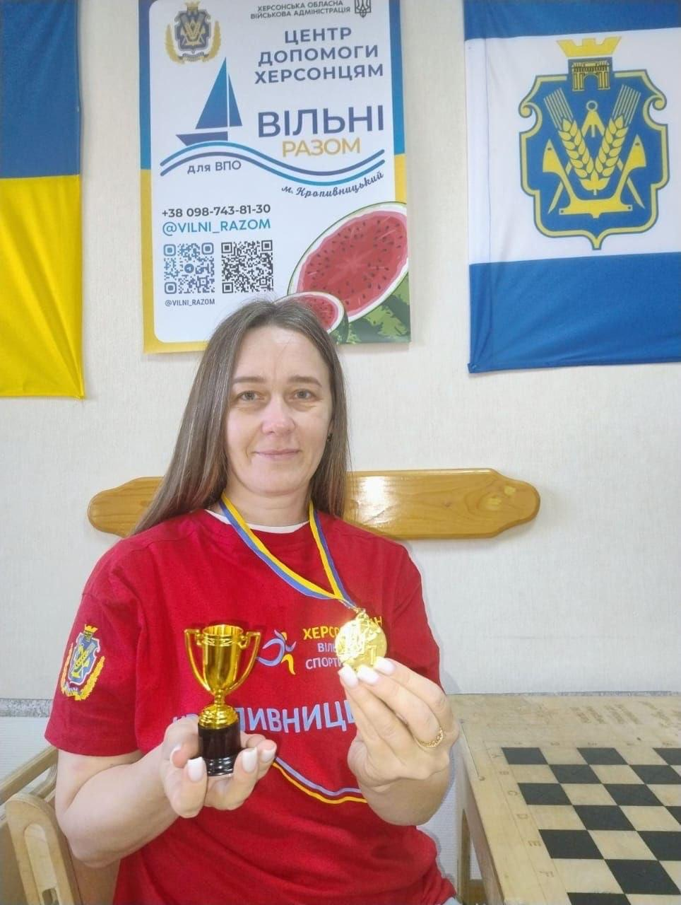 Жінка-переселенка з Херсонщини позує на тлі офіційної символіки хабу в Кропивницькому, тримаючи в руках завойовані нагороди — золотий кубок та медаль за перемогу в турнірі з шашок. Вона сидить перед великою дерев'яною ігровою дошкою, одягнена в патріотичну червону футболку з гербом та написом «Херсонщина. Вільні разом. Кропивницький». Жінка посміхається в кадр, тримаючи в правій руці маленький золотий кубок на чорній підставці, а в лівій — круглу золоту медаль на синьо-жовтій стрічці, яка одягнена їй на шию в межах фізкультурного заходу до Тижня спорту. На білій стіні заднього плану розвішано офіційні матеріали: ліворуч — Державний Прапор України, по центру — банер Центру допомоги херсонцям «Вільні разом» у м. Кропивницький із зображенням стиглої скибки кавуна та QR-кодами, а праворуч — біло-синій прапор Херсонської області з гербом.