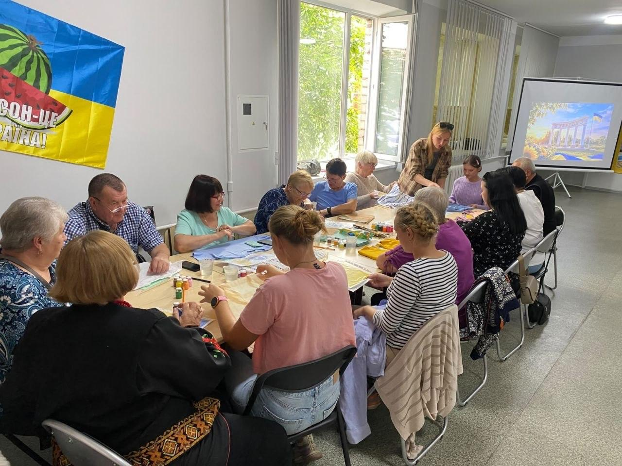 Група з п'ятнадцяти переселенців із Херсонщини та організаторів займається творчістю в житомирському координаційному центрі «Вільні разом» на урочистому заході з майстер-класом з розпису одягу до Дня Херсона. Чотирнадцятеро учасників різного віку розмістилися за довгим спільним столом, розписуючи елементи гардероба патріотичними малюнками за допомогою фарб і пензлів, а дівчина-модераторка у сорочці в клітинку допомагає їм, стоячи біля вікна. Ліворуч на стіні розгорнуто синьо-жовтий прапор із кавуном і гаслом «ХЕРСОН — ЦЕ УКРАЇНА!». На задньому плані в торці кімнати встановлено великий білий екран, на який через проєктор виведено яскраве кольорове зображення херсонського монумента на тлі сонячного пейзажу.