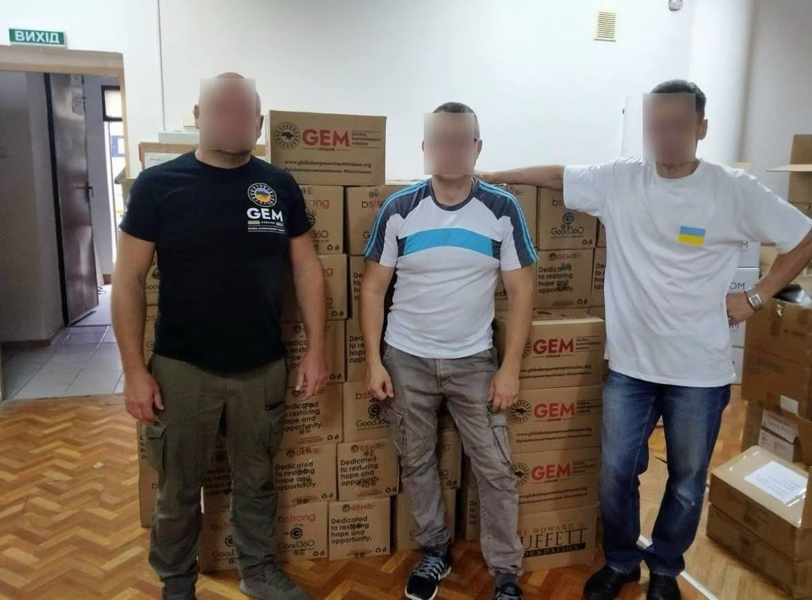 Три чоловіки із затушованими з міркувань безпеки обличчями стоять у приміщенні херсонського Центру гуманітарної допомоги Чулаківської СВА на тлі великої стіни з картонних коробок із продуктовими наборами від організації Global Empowerment Mission Ukraine (GEM) та фонду Howard G. Buffett Foundation (HGBF). Вони вишикувалися в один ряд перед високою барикадою з пакунків, що призначені для підтримки сімей з лівобережжя Херсонщини. Крайній ліворуч чоловік одягнений у чорну футболку з логотипом «GEM» та тактичні штани кольору хакі. По центру стоїть чоловік у світлій футболці із синьо-блакитними вставками на плечах, а крайній праворуч — у білій футболці з невеликим синьо-жовтим прапорцем на грудях та синіх джинсах, спершись рукою на коробки. На багатьох картонних ящиках позаду них чітко видно великі написи «GEM», «Good360» та логотипи благодійних фундацій, а ліворуч на задньому плані видно коридор із відкритими дверима та зеленим покажчиком з написом «ВИХІД».