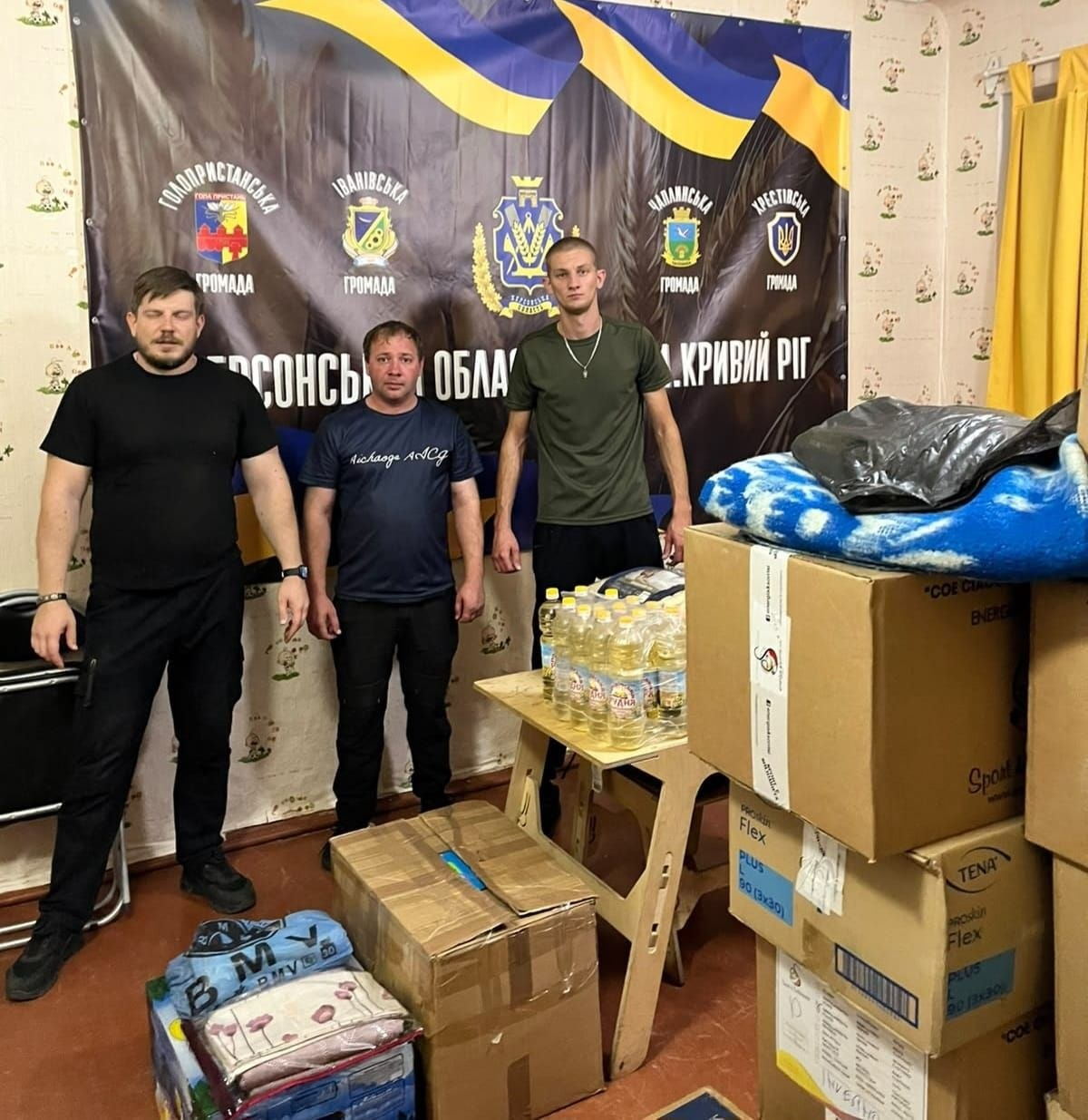 Група чоловіків стоїть у приміщенні криворізького хабу біля стосів картонних коробок та пакунків із гуманітарною допомогою, яку Голопристанська громада отримала від Славутської громади Хмельницької області в межах проєкту «Пліч-о-пліч: згуртовані громади». Три чоловіки вишикувалися в один ряд по центру кімнати. Чоловік ліворуч одягнений у чорну футболку, по центру стоїть чоловік у темно-синій футболці, а праворуч — високий хлопець у зеленій футболці. Перед ними на дерев'яному столі розставлені пляшки з соняшниковою олією, а поруч на стосах ящиків лежать пакунки з ковдрами, постільною білизною й памперсами для дорослих «TENA». На задньому плані височіє великий темний банер із написом «КРИВИЙ РІГ» та офіційними гербами Голопристанської, Іванівської, Чаплинської й Хрестівської територіальних громад під Державним Прапором України.
