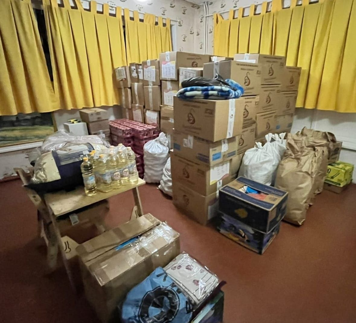 Приміщення складу хабу повністю заповнене високими стосами картонних коробок та великими мішками з гуманітарною допомогою, яку Голопристанська громада отримала від Славутської громади Хмельницької області у межах проєкту «Пліч-о-пліч: згуртовані громади». Пакунки та ящики з харчовими продуктами, одягом та засобами гігієни для ВПО акуратно складені вздовж стін кімнати під яскраво-жовтими віконними шторами. По центру височіє піраміда з коробок, на вершині якої лежить складена ковдра у синьо-білу клітинку. Ліворуч на дерев'яному столі розставлені пляшки з соняшниковою олією та упаковані текстильні вироби. Поруч на підлозі розміщені великі білі тканинні мішки та щільні паперові крафтові мішки, а у правому нижньому кутку стоїть відкрита коробка, де видно кольорові речі.