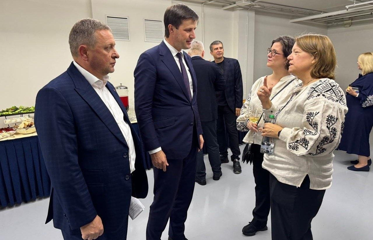 Перший заступник начальника Херсонської ОВА Дмитро Бутрій та Міністр аграрної політики та продовольства України Тарас Висоцький спілкуються з міжнародними партнерами під час зустрічі, присвяченої результатам відновлення аграрного сектору Херсонщини. На передньому плані ліворуч стоять двоє посадовців у класичних темно-синіх костюмах, обговорюючи питання підтримки українських фермерів у межах заходу, організованого міжнародною гуманітарною організацією Mercy Corps / Prosper Global. Навпроти них розмовляють дві жінки в білих вишиванках із чорним орнаментом, одна з яких тримає в руці пляшку води й активно жестикулює під час бесіди. На задньому плані у просторому світлому приміщенні розмовляють інші британські парламентарі та учасники зустрічі, а ліворуч уздовж стіни облаштовано фуршетний стіл із їжею та напоями.