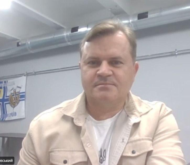 Заступник голови Херсонської ОДА Володимир Клюцевський у світло-бежевій вельветовій сорочці поверх білої футболки з гербом України проводить чергове засідання Гуманітарного штабу при Херсонській ОВА в режимі онлайн-трансляції. Посадовець перебуває по центру кадру на тлі світло-сірої стіни кабінету, під стелею якого проходить металева вентиляційна труба. Ліворуч на задньому плані розгорнуто прапор із символікою, а у лівому нижньому кутку екрана міститься плашка з білим написом його прізвища під час ухвалення рішень щодо забезпечення громад генераторами, пальним та засобами реабілітації для адаптивного спорту.