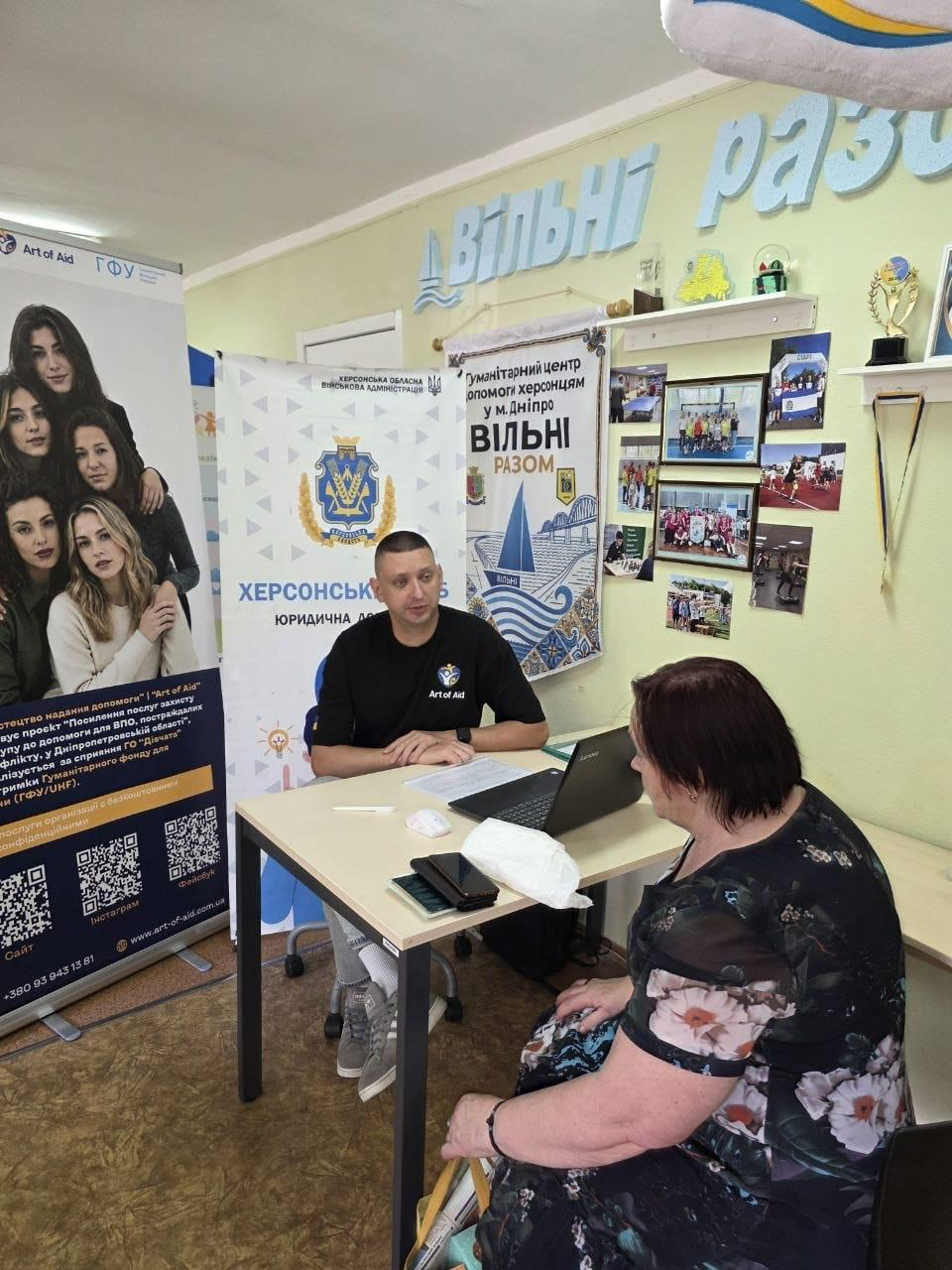 Кейс-менеджер благодійного фонду «GLOBA22» у чорній футболці проводить індивідуальну консультацію щодо соціального супроводу для відвідувачки в кабінеті дніпровського координаційного центру «Вільні разом». Фахівець сидить за світлим робочим столом біля відкритого ноутбука та роз'яснює переселенці з Херсонщини особливості отримання підтримки. Жінка з коротким темним волоссям у квітчастій сукні сидить навпроти боком до камери, слухаючи спеціаліста в межах серії індивідуальних прийомів для ВПО. Позаду працівника встановлено мобільний ролап-стенд юридичної допомоги з гербом Херсонської області, а на світло-зеленій стіні закріплено інформаційний плакат із написом «Гуманітарний центр допомоги херсонцям у м. Дніпро ВІЛЬНІ РАЗОМ» поруч із виставкою фотографій і нагородними кубками.