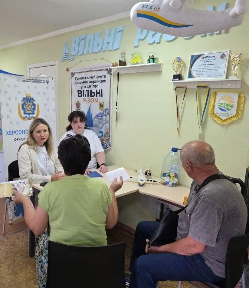 Юрисконсульти благодійних організацій надають індивідуальні консультації переселенцям із Херсонщини за кутовим робочим столом у приміщенні дніпровського координаційного центру «Вільні разом». Ліворуч за столом сидить юристка у світлому жакеті, поруч із якою перебуває інша фахівчиня у білій футболці. Навпроти них на стільцях розмістилися відвідувачі: жінка у світло-зеленій футболці, повернута спиною до кадру, та літній чоловік у сірій футболці й синіх джинсах. На задньому плані кабінету встановлено мобільний ролап-стенд юридичної допомоги з гербом Херсонської області, а на стіні закріплено плакат із написом «Гуманітарний центр допомоги херсонцям у м. Дніпро ВІЛЬНІ РАЗОМ» під великими об'ємними літерами. Також на стіні та поличках виставлено нагородні кубки, дипломи, символіку Великоолександрівської громади та м'яку іграшку у вигляді літака «Мрія UA».