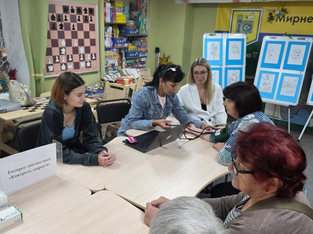 Медична працівниця у джинсовій куртці вимірює тиск відвідувачці у дніпровському координаційному центрі «Вільні разом» під час зустрічі із сімейним лікарем Центру первинної медико-санітарної допомоги Бахмутської районної ради. Захід проведено в межах Тижня фізичної культури та спорту для переселенців із Херсонщини. За робочим столом сидить лікар у білому піджаці та окулярах, яка надає консультації, виписує ліки та рекомендації, а поруч лежить відкритий ноутбук та встановлено табличку «Експрес-діагностика «Контроль здоров'я». Навколо столу розмістилися інші відвідувачки різного віку. На задньому плані кабінету на стіні закріплено демонстраційну шахову дошку, шафу з настільними іграми, сині стенди з малюнками та офіційний банер Мирненської громади.