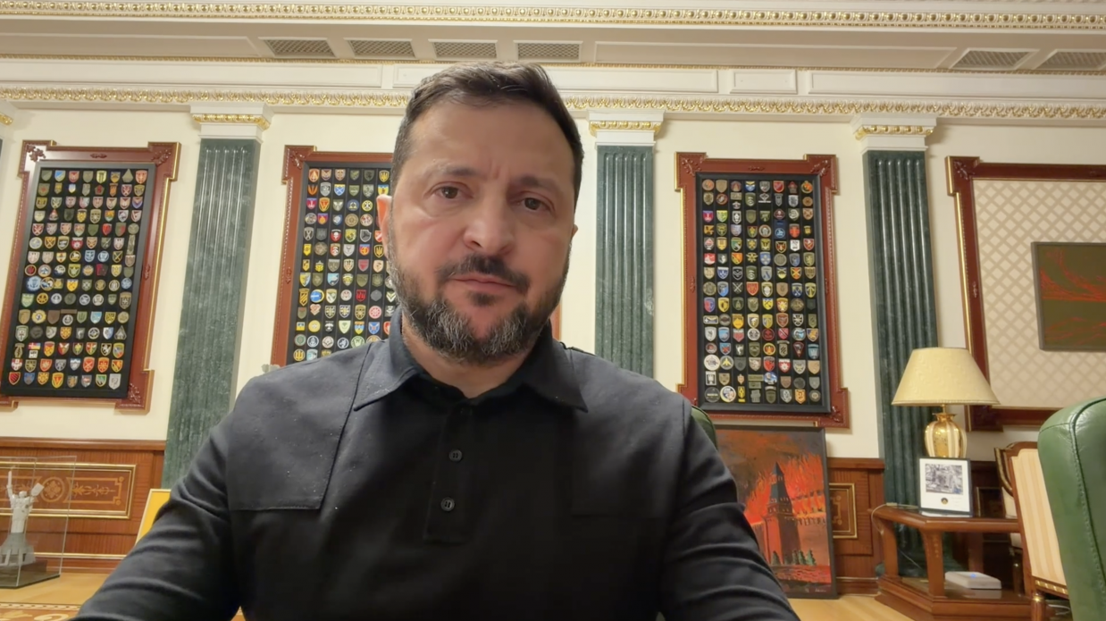 Президент України Володимир Зеленський у чорній сорочці-поло виступає з відеозверненням щодо підписання додаткових контрактів на системи захисту об’єктів Укрзалізниці та прикордонних переходів. Він перебуває по центру кадру в кабінеті на тлі стін із зеленими колонами та дошками з шевронами військових підрозділів.