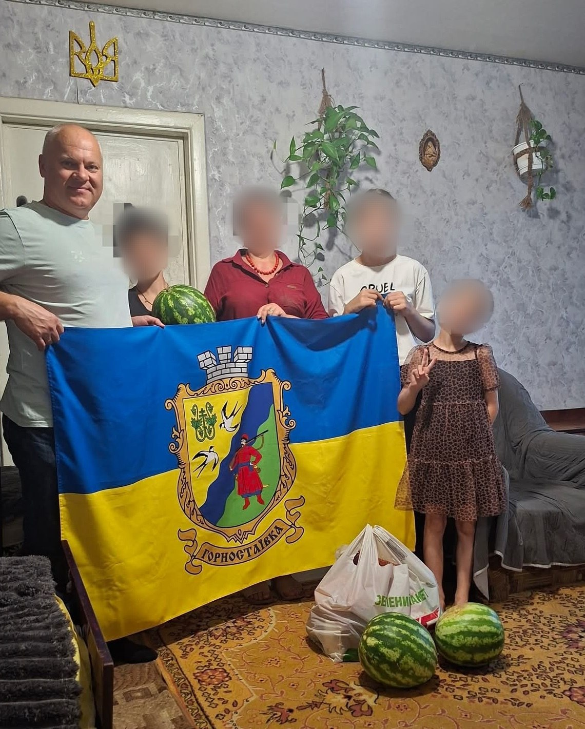 Начальник Горностаївської СВА Дмитро Ляхно відвідав родину переселенців у Дніпропетровській області в межах супроводу сімей із дітьми, яких вдалося повернути з тимчасово окупованих територій. З міркувань безпеки цивільного населення обличчя членів родини на фотографії затушовані, тоді як обличчя керівника громади залишається відкритим. Посадовець у світлій футболці стоїть ліворуч і разом із родиною тримає великий синьо-жовтий прапор Горностаївки з її офіційним гербом. Одна з учасниць зустрічі тримає великий стиглий кавун, а праворуч стоїть дівчинка у плямистій сукні, яка показує жест «вікторія». На передньому плані на килимі лежать ще два кавуни з правобережжя Херсонщини та стоїть пакет із солодкими подарунками, а на задньому плані над дверима закріплено золотавий Тризуб.