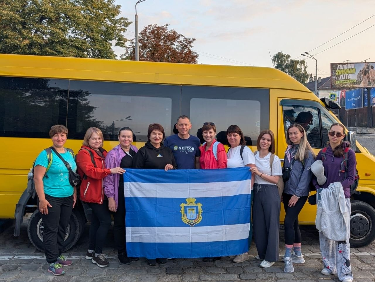 Група херсонців, які тимчасово проживають в Івано-Франківську, позує для спільного фото на тлі жовтого мікроавтобуса перед сходженням на Говерлу разом із благодійником Юрієм Рожковим, тримаючи великий синьо-білий прапор міста Херсона з його офіційним гербом. Учасники та учасниці заходу, організованого координаційним центром «Вільні разом», вишикувалися в один ряд на бруківці. По центру стоїть Юрій Рожков у темно-синій футболці з написом «ХЕРСОН», якого оточують жінки у спортивному та повсякденному одязі, серед яких члени родин військових, лікарі, вчителі та представниці ГО «Жінки обʼєднані війною». Усі присутні посміхаються й дивляться в об'єктив камери перед поїздкою до найвищої вершини України, щоб підняти там прапор до 248-річчя з дня заснування рідного міста.