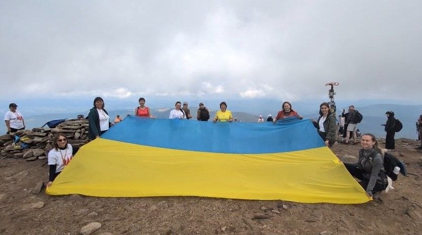 Група херсонців, які тимчасово проживають в Івано-Франківську, розгортає великий Державний Прапор України на вершині Говерли під час патріотичного заходу до 248-річчя з дня заснування рідного міста. Учасники та учасниці акції, організованої координаційним центром «Вільні разом» за фінансової підтримки Юрія Рожкова, вишикувалися на кам'янистому плато найвищої точки України. Вони тримають у руках величезне синьо-жовте полотнище, дивлячись в об'єктив камери та вигукуючи патріотичні гасла на тлі хмарного гірського неба Карпат, де серед присутніх на вершині зібралися лікарі, вчителі, студенти, члени родин військових та представниці ГО «Жінки обʼєднані війною».