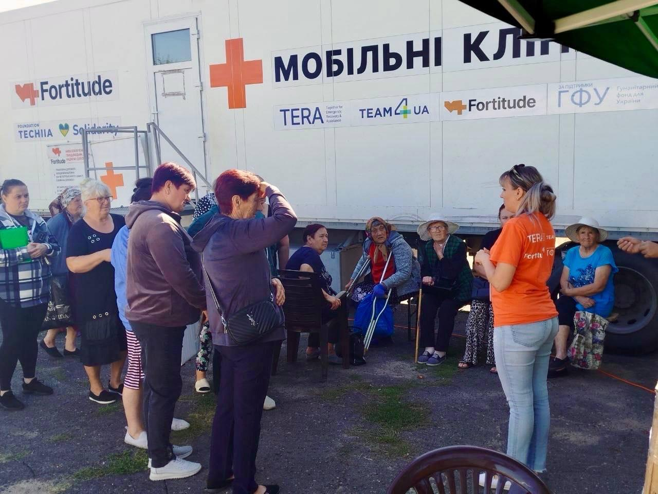 Група жителів Херсонщини очікує на прийом та слухає роз'яснення біля великого білого автомобіля мобільної клініки «Fortitude» під час виїзду медиків до Великоолександрівської та Високопільської громад. Праворуч на передньому плані стоїть представниця організації в яскравій помаранчевій футболці з фірмовими логотипами, яка спілкується з відвідувачами. Перед нею на стільчиках та в черзі розмістилися пацієнти переважно похилого віку, серед яких помітно жінку з милицями. На білому борту спеціалізованого комплексу нанесено великий напис «МОБІЛЬНІ КЛІНІКИ», червоний медичний хрест та логотипи партнерів, серед яких «Fortitude», «TERA», «TEAM4UA» та «ГФУ», де понад 450 мешканців сіл безоплатно отримували медикаменти та консультації профільних спеціалістів.