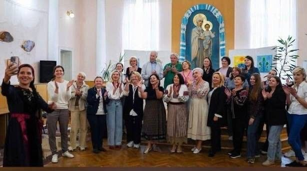 Група переселенців із Херсонщини та гостей спільного заходу позує для великого селфі із Заслуженою артисткою України Русланою Лоцман під час презентації родинного мистецького проєкту «Чари степового краю» майстрів із Херсонщини Інни Красновської-Кафтан та Миколи Кафтана в Івано-Франківську, організованої за сприяння координаційного центру «Вільні разом». На передньому плані ліворуч співачка в етнічному чорному вбранні з червоним поясом робить загальний знімок на мобільний телефон. Позаду неї півколом вишикувалися численні відвідувачі зустрічі у Навчально-методичному центрі культури і туризму Прикарпаття, які посміхаються та аплодують. На задньому плані світлої зали височіє велике настінне зображення Богородиці з немовлям в арковій блакитній рамі з орнаментами, а поруч помітно виставкові стенди з художніми роботами майстрів.