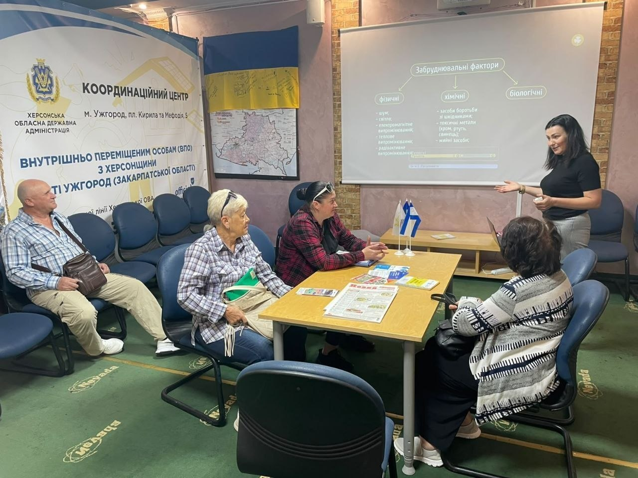 Представниця Міжнародного фонду охорони здоров'я та навколишнього середовища «Регіон Карпат» (NEEKA) у чорній футболці проводить правову консультацію для переселенців із Херсонщини в ужгородському координаційному центрі «Вільні разом». Вона стоїть праворуч біля великого білого проєкційного екрана, на який виведено слайд із заголовком «Забруднювальні фактори» та блоками про фізичні, хімічні й біологічні чинники. Навколо столу по центру та ліворуч розмістилися слухачі, які прийшли отримати юридичну допомогу. На столі перед ними лежать інформаційні друковані матеріали, брошури та встановлено малий прапорець України, а на стіні позаду закріплено карту та офіційний банер «Координаційний центр Херсонської обласної державної адміністрації м. Ужгород» під великим Державним Прапором.