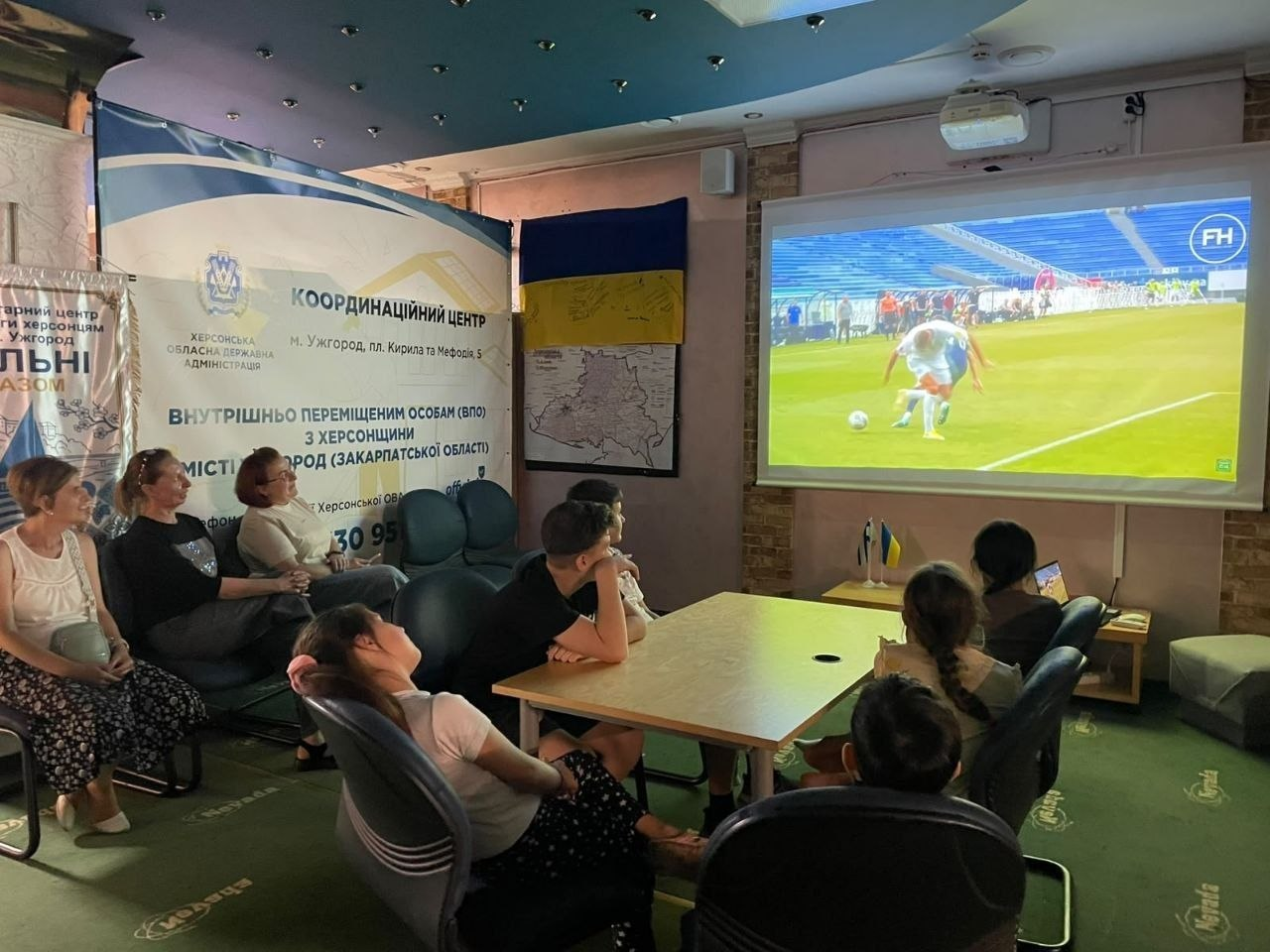 Переселенці з Херсонщини різного віку сидять у залі ужгородського координаційного центру «Вільні разом», уважно переглядаючи фільм «Фокс і Макс» на великому білому екрані кіноклубу «Кіно без кордонів» з нагоди Дня українського кіно. Глядачі розмістилися ліворуч на офісних стільцях та чорному шкіряному дивані, а також навколо дерев'яного столу по центру, повернувшись обличчям до стіни з мультимедійним проєктором, де 14 ВПО долучилися до культурного дозвілля. На великому спроєктованому екрані демонструється кадр із фільму із футболістом у білій формі на тлі синіх трибун стадіону. Позаду присутніх ліворуч встановлено великий офіційний банер «Координаційний центр Херсонської обласної державної адміністрації м. Ужгород», а поруч на стіні під Державним Прапором України закріплено контурну карту Херсонської області.