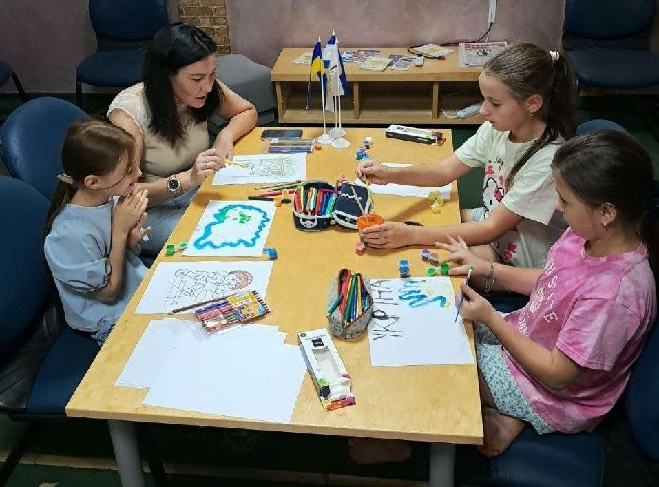 Діти переселенців із Херсонщини разом із дорослою наставницею малюють за дерев'яним столом в ужгородському координаційному центрі «Вільні разом» під час конкурсу дитячого малюнка «Мій улюблений вид спорту», організованого на базі дитячого артмайданчика. Ліворуч сидить жінка у світлому топі, яка допомагає дівчинці в блакитній футболці розфарбовувати малюнки олівцями. Праворуч за столом розмістилися ще дві дівчинки: одна у білій футболці відкриває баночку з фарбою, а інша в рожевій футболці малює пензликом на аркуші поруч із патріотичним написом «УКРАЇНА». На столі між учасниками розкладено велику кількість кольорових олівців, фломастерів, баночок із гуашшю, розмальовок, а по центру встановлено два маленькі настільні прапорці — України та міста Херсона.