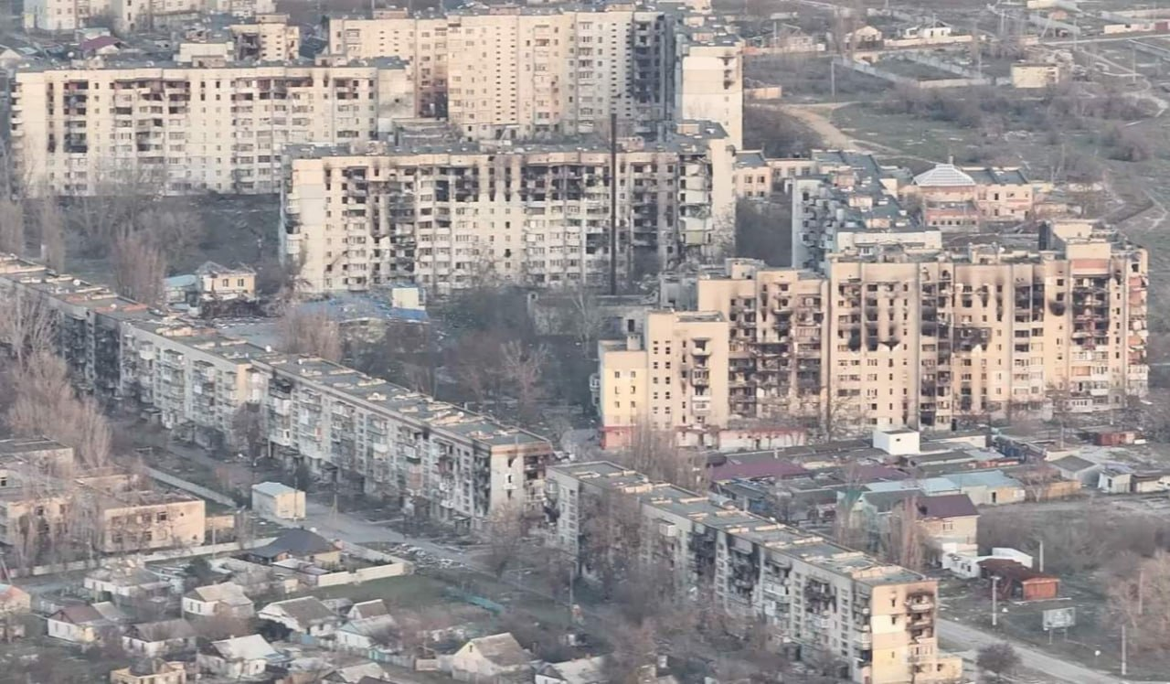 Вид з висоти пташиного польоту на зруйновані внаслідок російської агресії житлові квартали міста Олешки на Херсонщині. На знімку зафіксовано масштабні пошкодження багатоповерхових панельних будинків цивільної інфраструктури. На фасадах будівель чітко видно чорні сліди від пожеж після обстрілів, вибиті вікна, понівечені балкони та зруйновані міжповерхові перекриття окремих квартир. Навколо багатоповерхівок розташована приватна забудова, пустирі та поодинокі дерева без листя. Панорама демонструє наслідки бойових дій та гуманітарної катастрофи в заблокованому місті.