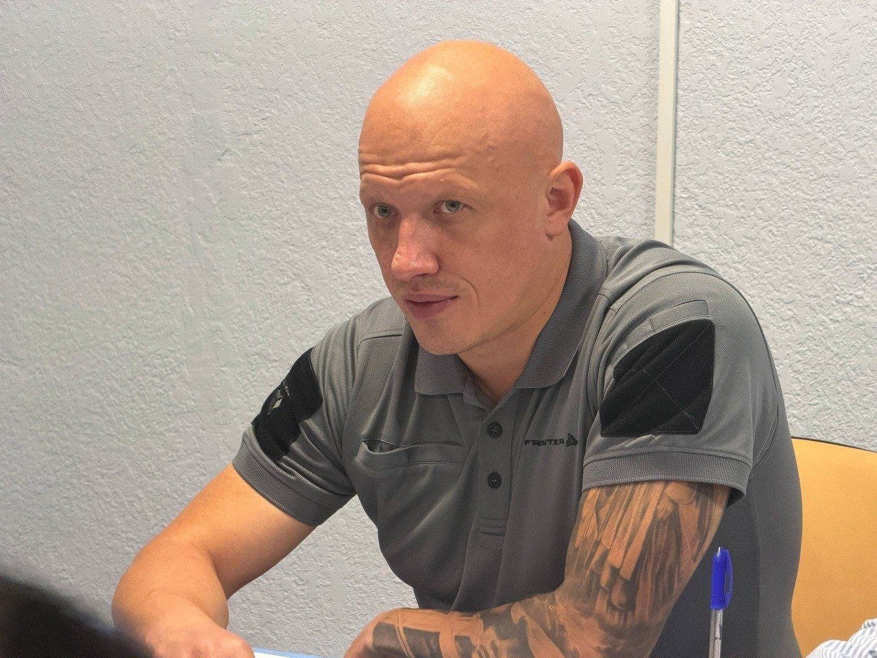 Заступник начальника Херсонської ОВА Євгеній Ігнатенко у сірій футболці-поло сидить за робочим столом у кабінеті під час робочої зустрічі з делегацією Міжнародного Комітету Червоного Хреста у сфері посилення медичної системи та безпеки лікарень регіону. Керівник зображений крупним планом до пояса, повернутий напівпрофілем ліворуч і уважно слухає співрозмовників, обговорюючи підготовку лікарень до осінньо-зимового періоду та захист медичних працівників від російських обстрілів. На рукавах його футболки помітні чорні тактичні велкро-панелі, а на столі перед ним стоїть синя кулькова ручка на тлі світлої фактурної стіни приміщення.