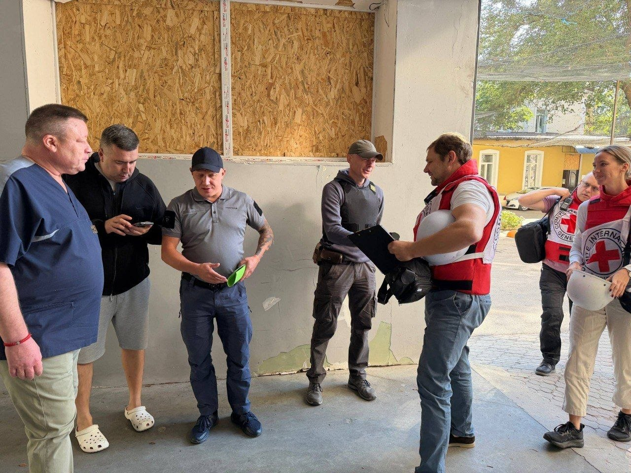 Група чоловіків та представників Міжнародного Комітету Червоного Хреста у фірмових червоно-білих жилетах і накидках із великими логотипами організації спілкується на подвір'ї медичного закладу в межах системної співпраці щодо посилення медичної системи та безпеки лікарень Херсонщини. Ліворуч стоїть Генеральний директор обласної клінічної лікарні Віктор Короленко у темно-синьому медичному костюмі, поруч із ним чоловік у чорній кофті дивиться у мобільний телефон, а по центру заступник начальника Херсонської ОВА Євгеній Ігнатенко у сірій футболці-поло та синій кепці тримає в руках зелений блокнот. Навпроти них представник МКЧХ у червоному тактичному жилеті робить записи у робочому планшеті для фіксації потреб установи напередодні нового опалювального сезону, тоді як праворуч за ними спостерігають двоє колег гуманітарної місії. Зустріч відбувається на тлі бетонної стіни з віконним отвором, закритим плитою ОСП, а праворуч у прорізі видно сонячну вулицю та сусідню будівлю.
