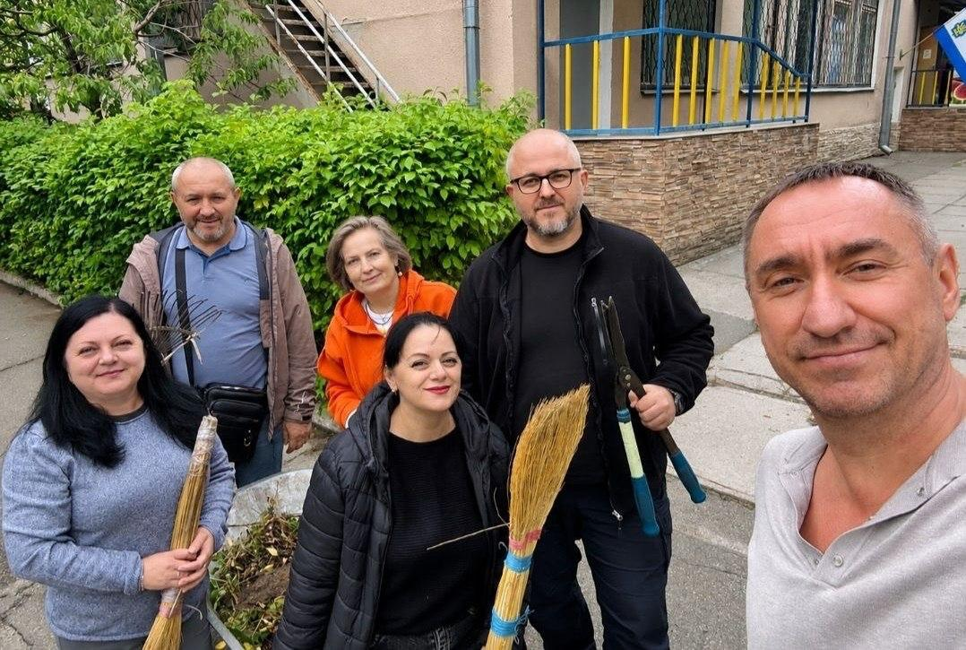 Група представників київського координаційного центру «Вільні разом» та переселенців із Херсонщини робить спільне селфі біля будівлі простору під час толоки «Разом за чисте довкілля» до Всесвітнього дня прибирання. На передньому плані праворуч чоловік у світлій футболці-поло тримає телефон і робить загальний знімок, а навколо нього зібралися колеги з господарським інвентарем: чоловік у чорній кофті та окулярах тримає великі садові ножиці, а ліворуч дві жінки тримають віники з сорго. На задньому плані на тлі зелених кущів посміхаються чоловік із граблями в руках та жінка в яскраво-помаранчевій кофті, а праворуч зверху на краю фото помітно на будівлі частково прапор Херсонської області.