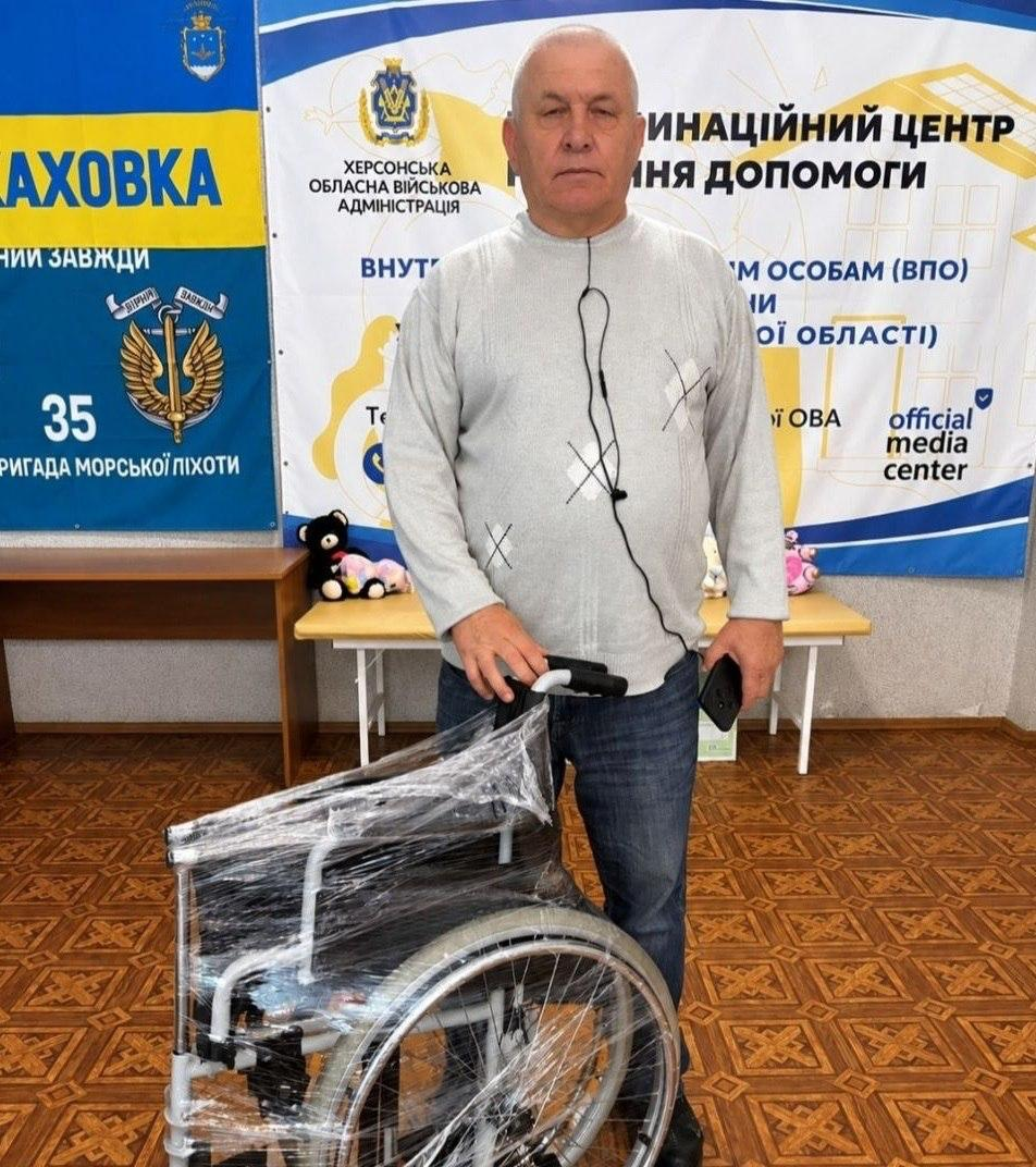 Чоловік старшого віку у сірій кофті в клітинку та синіх джинсах стоїть по центру залу координаційного центру, тримаючи правою рукою ручку нового інвалідного візка, який обгорнутий захисною прозорою плівкою. Чоловік зображений у повний зріст, повернутий обличчям до камери, а в лівій руці тримає мобільний телефон. Позаду нього на стіні розвішано офіційні банери: ліворуч синьо-жовтий плакат із написом «КАХОВКА» та символікою 35-ї окремої бригади морської піхоти, а праворуч — білий банер із гербом Херсонської обласної військової адміністрації та написами «КООРДИНАЦІЙНИЙ ЦЕНТР» та «official media center». На задньому плані між банерами видно дерев'яний стіл, на якому лежать м'які дитячі іграшки, а підлога в приміщенні вистелена коричневим лінолеумом із геометричним візерунком.