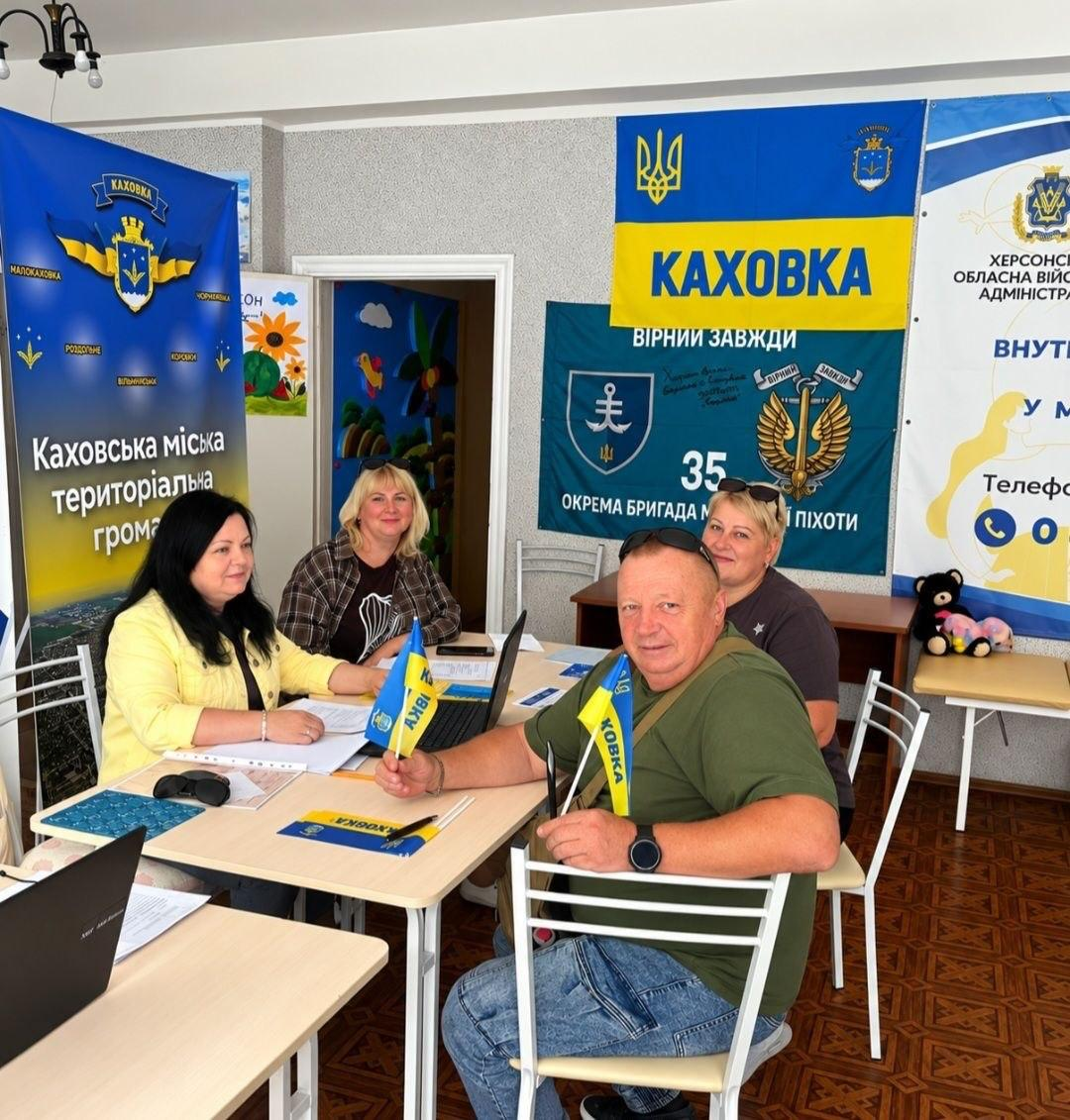 Група переселенців із Херсонщини сидить за столами в київському координаційному центрі «Вільні разом» під час організації благодійного Фестивалю єдності «Херсонщина - символ нескореності» зі збору коштів на детектор дронів «Чуйка 4.0» для 34 ОБрБО. Ліворуч дві жінки працюють із документами, а праворуч на стільці сидить чоловік у зеленій футболці, який тримає в руці маленький прапорець із написом «КАХОВКА», на тлі ще однієї відвідувачки. На стінах зали розвішано офіційний стенд Каховської громади, Державний Прапор України з написом «КАХОВКА / ВІРНІ ЗАВЖДИ», темно-синій прапор 35-ї окремої бригади морської піхоти та білий банер із гербом Херсонської області.