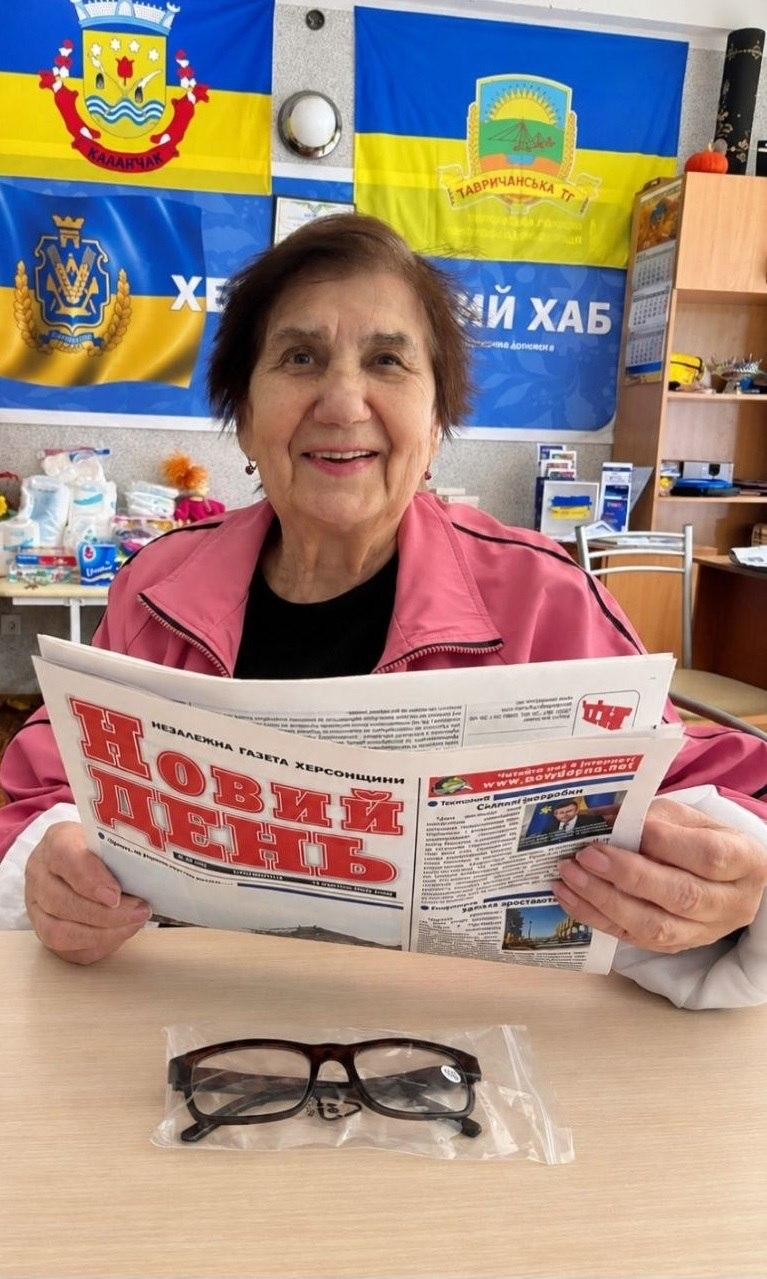 Літня жінка у рожевій куртці посміхається за столом у київському координаційному центрі «Вільні разом» під час видачі 55 пар окулярів для читання переселенцям поважного віку з Херсонщини. Вона тримає в руках газету «Новий день», а перед нею лежать окуляри у прозорому чохлі на тлі розвішаних на стіні прапорів міста Каховка, Тавричанської громади, 35-ї окремої бригади морської піхоти та банера «ХЕРСОНСЬКИЙ ХАБ».
