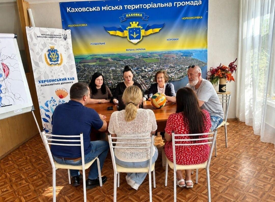 Група переселенців із Херсонщини та відомих спортсменів сидить навколо великого дерев'яного столу в залі київського координаційного центру «Вільні разом» під час круглого столу «Спорт, що об’єднує», організованого до Тижня спорту в межах всебічної підтримки внутрішньо переміщених осіб. На задньому плані за столом обличчям до камери сидять три жінки, а праворуч від них — літній сивобородий чоловік у сірій футболці, який тримає на столі волейбольний м'яч, тоді як на передньому плані спиною до кадру розмістилися чоловік у синій футболці, дівчина у світлій ажурній кофті та дівчина в червоній сукні. Зустріч, на якій обговорюють розвиток спорту, проходить на тлі великого офіційного банера «Каховська міська територіальна громада» із зображенням герба міста та мобільного стенда «Херсонський хаб / психологічна допомога» з патріотичним орнаментом.