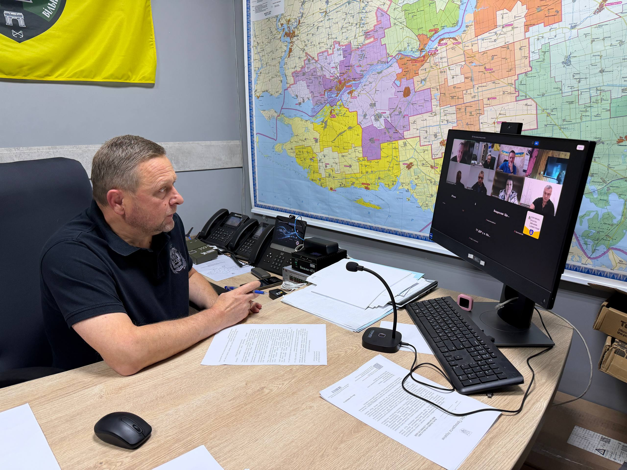 Перший заступник начальника Херсонської ОВА Дмитро Бутрій у темно-синій футболці-поло сидить за робочим столом у кабінеті під час чергового засідання обласної міжвідомчої робочої групи «Прозорість і підзвітність» щодо стану будівництва бомбосховищ та захисних споруд у навчальних закладах Бериславського району. Керівник зображений напівпрофілем праворуч та уважно дивиться на великий монітор комп'ютера з вікнами учасників відеоконференції, з якими обговорює суворий контроль за витратами бюджетних коштів у громадах. На столі перед ним розкладено документи, робочі папери, клавіатуру, комп'ютерну мишу, настільний мікрофон та стаціонарні телефони. На стіні позаду монітора розміщено велику детальну карту Херсонської області з різнокольоровими зонами громад, а ліворуч угорі помітно частину жовтого прапора з логотипом координаційного центру «Вільні разом».