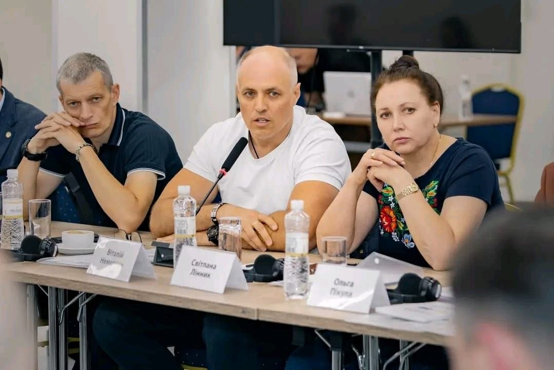 Учасники засідання круглого столу в Києві щодо роботи Міжнародного реєстру збитків, завданих агресією російської федерації проти України, зокрема за участю делегатів від Каховської, Голопристанської та Олешківської громад Херсонщини сидять за робочим столом і уважно слухають доповідачів. По центру обличчям до камери у білій футболці сидить начальник Каховської МВА Віталій Немерець, перед яким встановлено настільний мікрофон, праворуч від нього — начальниця Голопристанської МВА Світлана Лінник у темно-синій блузі з яскравим квітковим принтом, а ліворуч — чоловік у темно-синій футболці-поло, який підпер рукою підборіддя, обговорюючи алгоритми залучення та верифікації даних від постраждалих фізичних і юридичних осіб. На столі перед ними розставлені пляшки з мінеральною водою, склянки, навушники для синхронного перекладу, розкладені робочі папери та настільні таблички з іменами учасників на тлі великого чорного екрана.