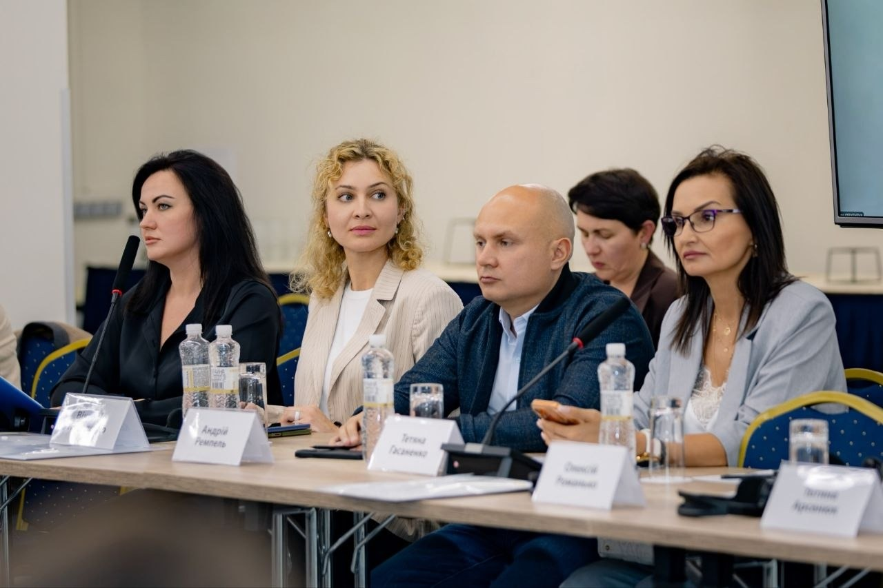 Учасники засідання круглого столу в Києві щодо роботи Міжнародного реєстру збитків, завданих агресією російської федерації проти України, зокрема за участю делегатів від Каховської, Голопристанської та Олешківської громад Херсонщини сидять за довгим робочим столом і уважно слухають виступи колег, обговорюючи алгоритми верифікації даних постраждалих осіб. За столом зліва направо розмістилися чотири людини: жінка-брюнетка у чорній сорочці, перед якою стоїть табличка з іменем Андрій Реутець, начальниця Олешківської МВА Тетяна Гасаненко у світлому жакеті, чоловік у темно-синій кофті на блискавці з табличкою Олексій Ромашко та жінка в окулярах і сірому піджаку з табличкою Тетяна Лук'янова. На столі перед ними розставлені пляшки з водою, склянки, настільні мікрофони для виступів та білі картки учасників засідання на тлі світлої стіни конференц-залу.