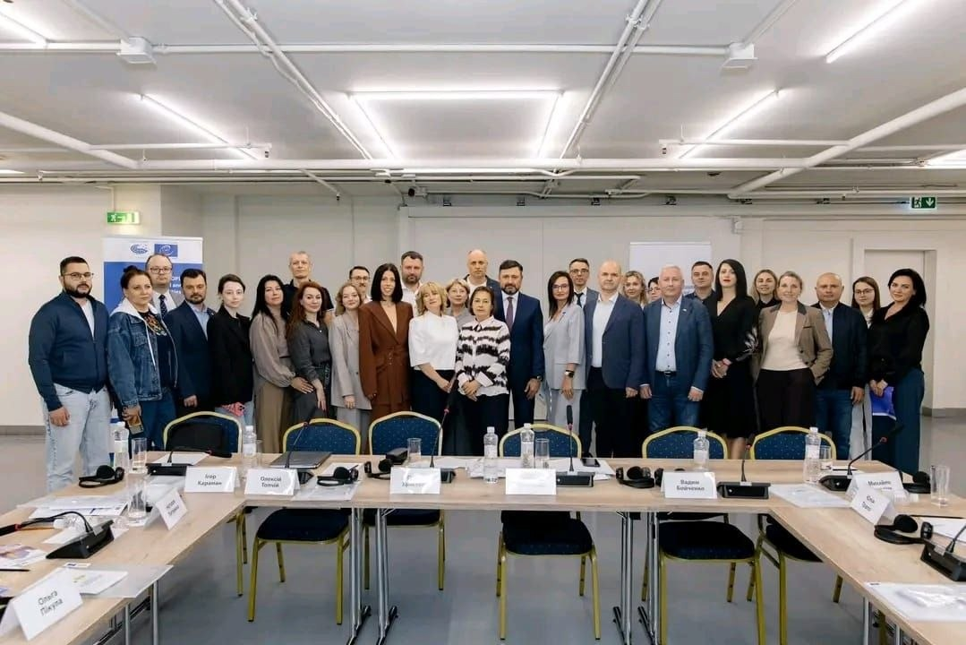 Велика група учасників засідання круглого столу в Києві щодо роботи Міжнародного реєстру збитків, завданих агресією російської федерації проти України, зокрема за участю делегатів від Каховської, Голопристанської та Олешківської громад Херсонщини позує для спільного фото в конференц-залі. Понад тридцять представників місцевих військових адміністрацій та міжнародних інституцій стоять у кілька рядів по центру залу для фіксації підсумків обговорення алгоритмів верифікації даних постраждалих осіб. На передньому плані перед ними розташовані довгі столи, розставлені літерою «П», на яких розміщені настільні мікрофони, навушники для перекладу, пляшки з мінеральною водою та білі картки з іменами учасників на тлі світлих стін та інформаційного банера Ради Європи ліворуч.