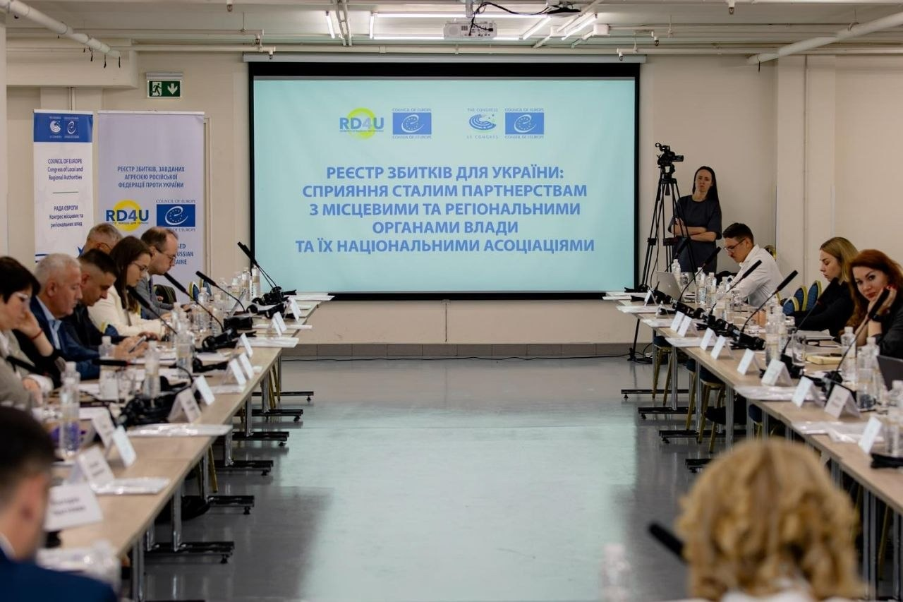 Учасники засідання круглого столу в Києві щодо роботи Міжнародного реєстру збитків, завданих агресією російської федерації проти України, зокрема за участю делегатів від Каховської, Голопристанської та Олешківської громад Херсонщини сидять за довгими столами в конференц-залі. По центру на великому екрані відображено головну тему зустрічі: «РЕЄСТР ЗБИТКІВ ДЛЯ УКРАЇНИ: СПРИЯННЯ СТАЛИМ ПАРТНЕРСТВАМ З МІСЦЕВИМИ ТА РЕГІОНАЛЬНИМИ ОРГАНАМИ ВЛАДИ ТА ЇХ НАЦІОНАЛЬНИМИ АСОЦІАЦІЯМИ» з логотипами Ради Європи та абревіатурою RD4U, які також продубльовані на білих банерах ліворуч. Присутні обговорюють алгоритми верифікації даних постраждалих осіб та специфіку руйнувань на окупованих територіях на тлі розставлених на столах мікрофонів, пляшок із водою, табличок та дівчини-оператора з відеокамерою на штативі праворуч.