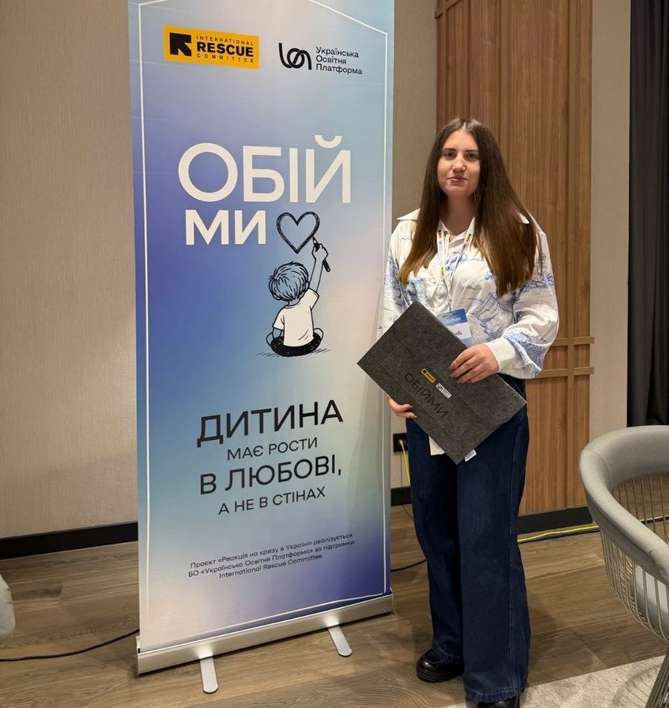 Начальниця служби у справах дітей Верхньорогачицької селищної ради Херсонщини стоїть у повний зріст у приміщенні під час навчання «Зустріч громад: там, де починається родина» з розвитку сімейних форм виховання. Вона одягнена у світлу блузу з блакитним візерунком та широкі сині джинси, а в руках тримає темно-сіру фетрову папку з написом «ОБІЙМИ». Ліворуч від неї встановлено вертикальний інформаційний банер проєкту «ОБІЙМИ» від International Rescue Committee та БО «Українська Освітня Платформа» з малюнком дитини, яка малює серце, та великим гаслом: «ДИТИНА МАЄ РОСТИ В ЛЮБОВІ, А НЕ В СТІНАХ» на тлі світлого залу з ламінатом та дерев'яними панелями.