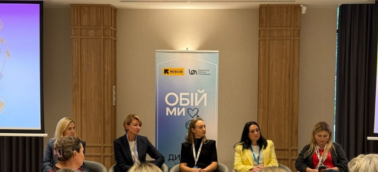 П'ять жінок-спікерок сидять у кріслах на тлі інформаційного банера під час панельної дискусії на навчанні «Зустріч громад: там, де починається родина» з розвитку сімейних форм виховання, у якому взяла участь начальниця служби у справах дітей Верхньорогачицької селищної ради Херсонщини. Учасниці президії повернуті обличчям до залу та слухають виступи колег, обговорюючи забезпечення права дитини на родину й інструменти підтримки фахівців соціальної сфери, а всі інші учасники заходу сидять на передньому плані спиною до камери та слухають доповідачок. По центру позаду спікерок встановлено вертикальний блакитно-білий стенд проєкту «ОБІЙМИ» з логотипами International Rescue Committee та БО «Українська Освітня Платформа», ліворуч помітно великий мультимедійний екран, а стіни кімнати декоровані вертикальними дерев'яними панелями.