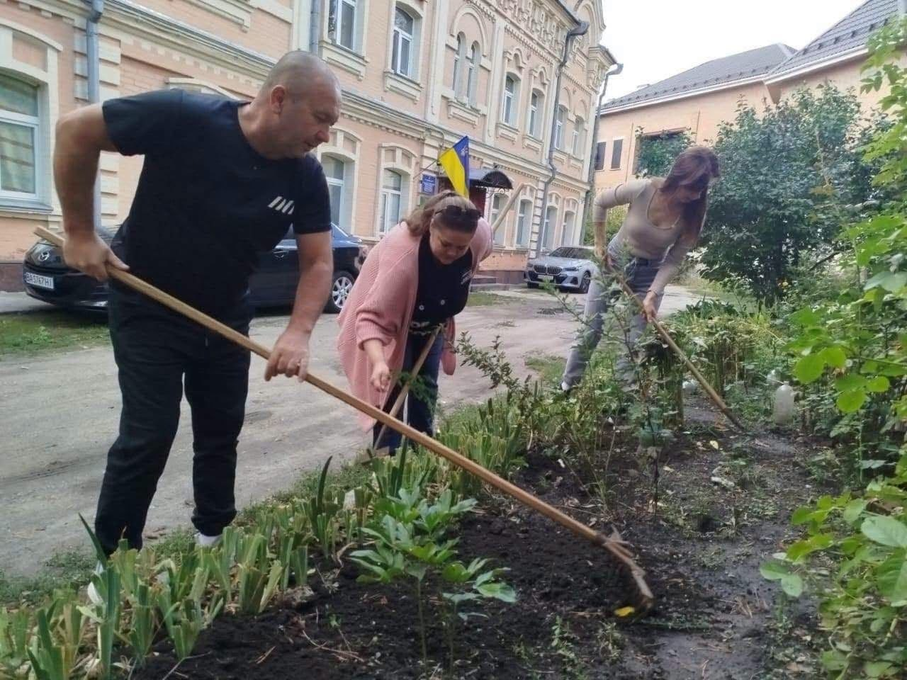 Працівники Верхньорогачицької СВА та переселенці з громади впорядковують клумбу перед історичною двоповерховою будівлею з жовтої цегли, долучившись до всеукраїнської екологічної ініціативи «World Cleanup Day 2026» до Всесвітнього дня прибирання. На передньому плані ліворуч чоловік у чорній футболці розпушує землю граблями, поруч із ним по центру схилилася жінка у рожевому кардигані, яка упорядковує зелені насадження, а праворуч інша дівчина за допомогою сапи обробляє ґрунт серед кущів, приділяючи увагу екологічному вихованню. На задньому плані на тлі цегляного фасаду будівлі майорить невеликий Державний Прапор України над входом, а на узбіччі припарковані легкові автомобілі.