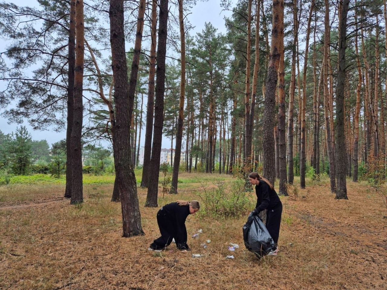 Дві дівчини у чорному спортивному одязі з числа переселенців Верхньорогачицької громади збирають розкидане сміття у хвойному лісі, долучившись до всеукраїнської екологічної ініціативи «World Cleanup Day 2026» до Всесвітнього дня прибирання. Одна з учасниць нахилилася, піднімаючи з сухої глиці та трави пластикові пляшки й папір, а інша тримає великий чорний поліетиленовий пакет для збору та подальшого сортування відходів на тлі високих сосен, зелених кущів та обрисів багатоповерхових будинків далеко на задньому плані.