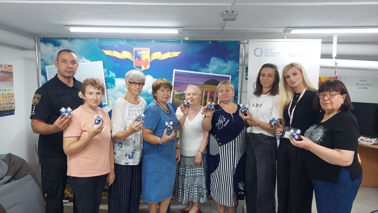 Велика група людей, що складається з чоловіка у чорній формі поліції та восьми жінок різного віку, стоїть у ряд у світлому приміщенні хабу й посміхається в кадр, демонструючи в руках невеликі хендмейд-вироби, а на задньому плані за ними встановлено великий яскравий банер із зображенням герба Голої Пристані та краєвидів міста. Заняття організовано для психоемоційного відновлення та підтримки громадян, оскільки протягом тижня у Центрі життєстійкості проводились арттерапевтичні та інформаційні заходи, зокрема відбувся творчий майстер-клас «Сова», де учасники власноруч виготовляли сувеніри, спілкувалися та отримували корисну психосоціальну допомогу.