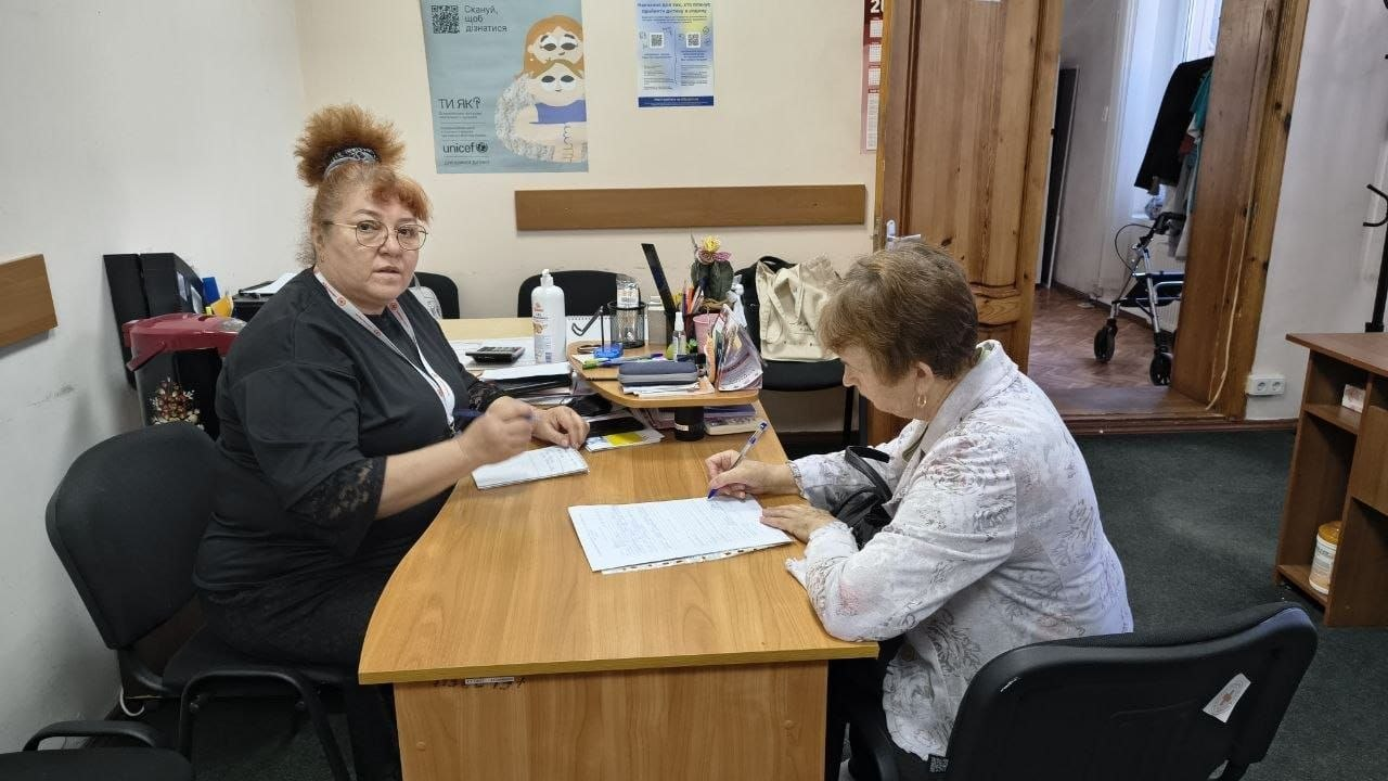 Жінка-фахівчиня в окулярах та чорній кофті сидить за дерев'яним кутовим столом у кабінеті й веде прийом літньої жінки у світлій блузі з візерунком, яка сидить навпроти та ручкою заповнює паперовий бланк. На столі розкладено канцелярію, а на стіні позаду висять плакати Всеукраїнської програми ментального здоров'я «Ти як?» від ЮНІСЕФ. Надання індивідуальної підтримки організовано для всебічної допомоги громадянам, оскільки протягом тижня у Центрі життєстійкості проводились арттерапевтичні та інформаційні заходи, зокрема проводилась індивідуальна робота з відвідувачами задля надання психосоціальної підтримки, консультування щодо отримання соціальних послуг та покращення ментального здоров'я родин.