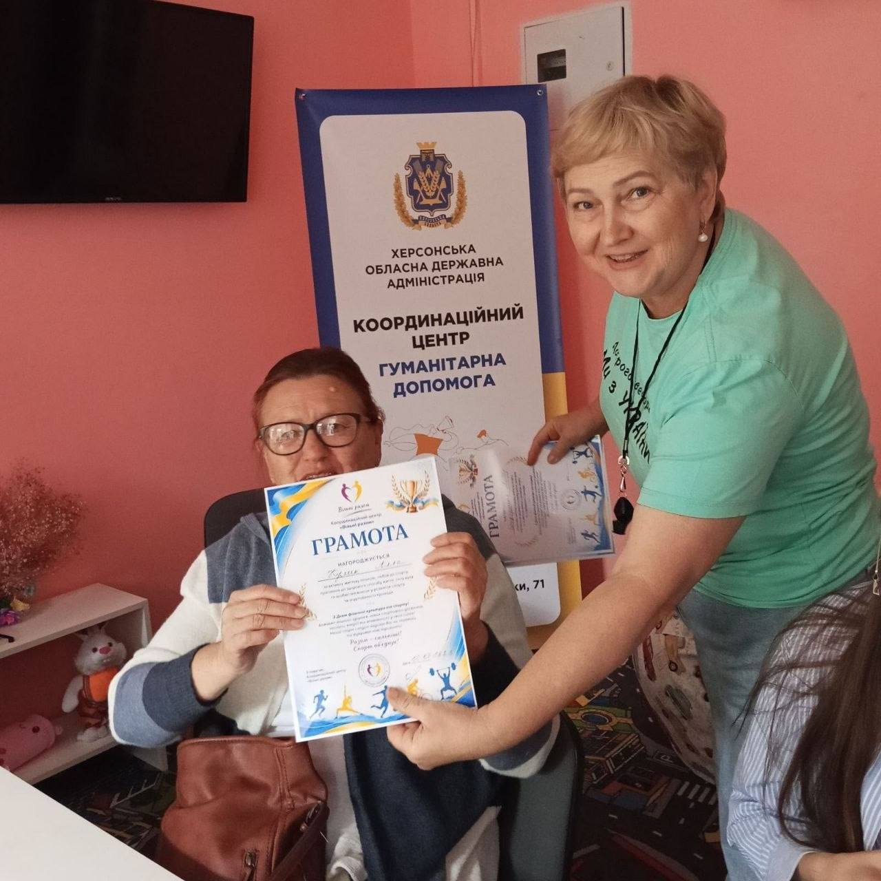 Жінка в окулярах сидить у кріслі й демонструє грамоту з малюнками спортсменів, а поруч стоїть працівниця хабу з іншою грамотою на тлі банера Херсонської ОДА з написом КООРДИНАЦІЙНИЙ ЦЕНТР ГУМАНІТАРНА ДОПОМОГА. Активності організовано для внутрішньо переміщених осіб за сприяння координаційного центру Вільні разом у Львові, де у межах Тижня фізичної культури та спорту та Дня турботи про здоров’я відбулись інтерактивний захід, експрес-діагностика «Контроль твого здоров’я» та практична зустріч «Займатися спортом чи робити зарядку?» задля просування здорового способу життя та психоемоційного відновлення родин.