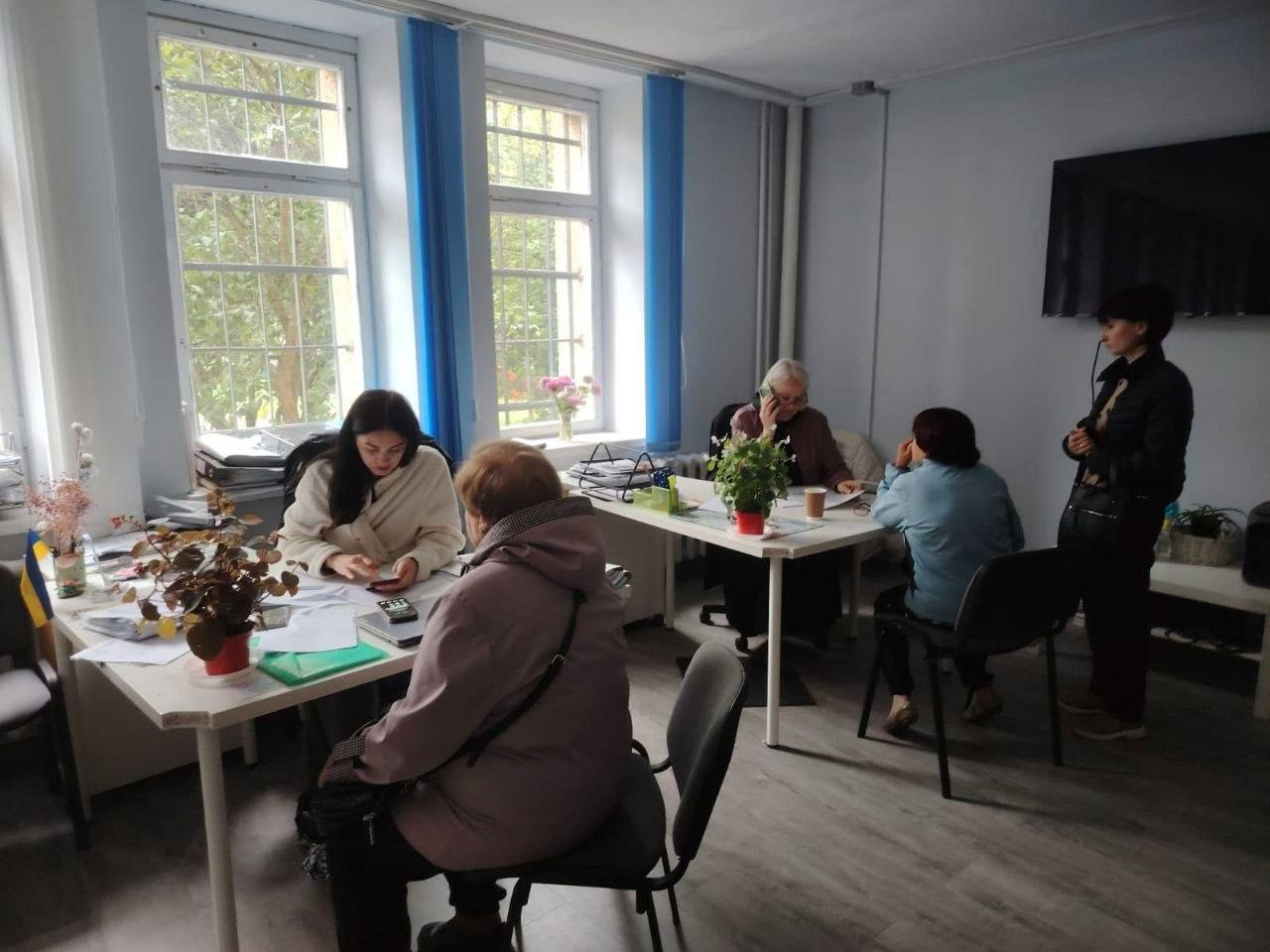 Робоча атмосфера в кабінеті хабу, де за двома білими столами фахівці ведуть прийом громадян на тлі великих вікон. Ліворуч молода працівниця консультує літню жінку в бузковій куртці, а на задньому плані інша консультантка розмовляє по телефону, приймаючи відвідувачку у блакитній сорочці. Робота організована в межах діяльності координаційного центру Вільні разом у Львові, фахівці якого системно підтримують переселенців з Херсонщини та надавали індивідуальні консультації з питань отримання адміністративних, соціальних, юридичних послуг, а також допомоги у працевлаштуванні внутрішньо переміщених осіб.
