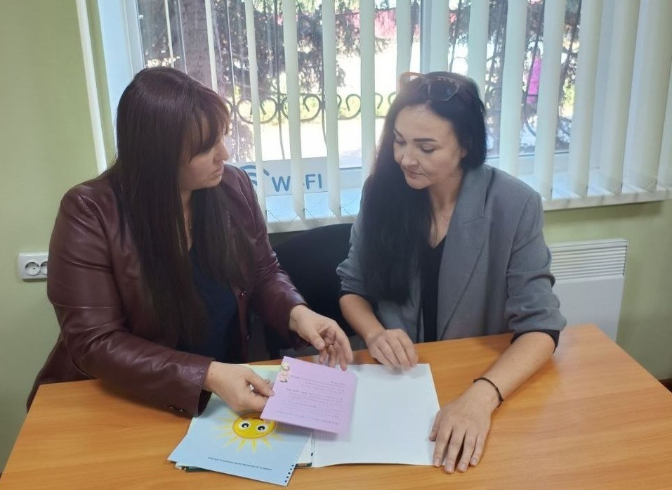 Фахівчиня служби у справах дітей Музиківської громади в бордовій шкіряній куртці та жінка у сірому піджаку сидять за столом у кабінеті й вивчають буклет із малюнком сонця та інформаційні листівки. Зустріч присвячена соціальній підтримці, оскільки фахівці служби у справах дітей Музиківської громади провели інформаційно-роз'яснювальні бесіди з жителями щодо популяризації сімейних форм виховання дітей, зокрема усиновлення, опіки та піклування, прийомних сімей задля ознайомлення кандидатів із порядком набуття статусу та умовами виховання малечі в межах заходів до Дня усиновлення, які відбулись у громадах Херсонщини.