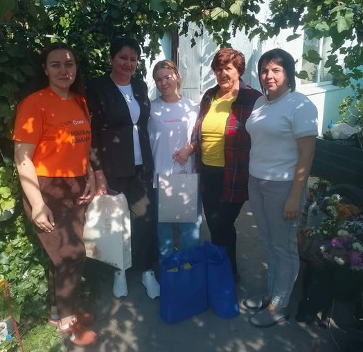 П'ять жінок позують для спільного фото на подвір'ї приватного будинку в оточенні зеленого виноградника, тримаючи в руках та біля ніг великі сині сумки й паперові пакети з речами. Благодійний візит організовано з нагоди свята, оскільки представники Великоолександрівської селищної ради та БФ «Вітри змін» відвідали прийомну сім’ю громади та вручили їм подарунки, продемонструвавши підтримку та турботу в межах заходів до Дня усиновлення, які відбулись у громадах Херсонщини.