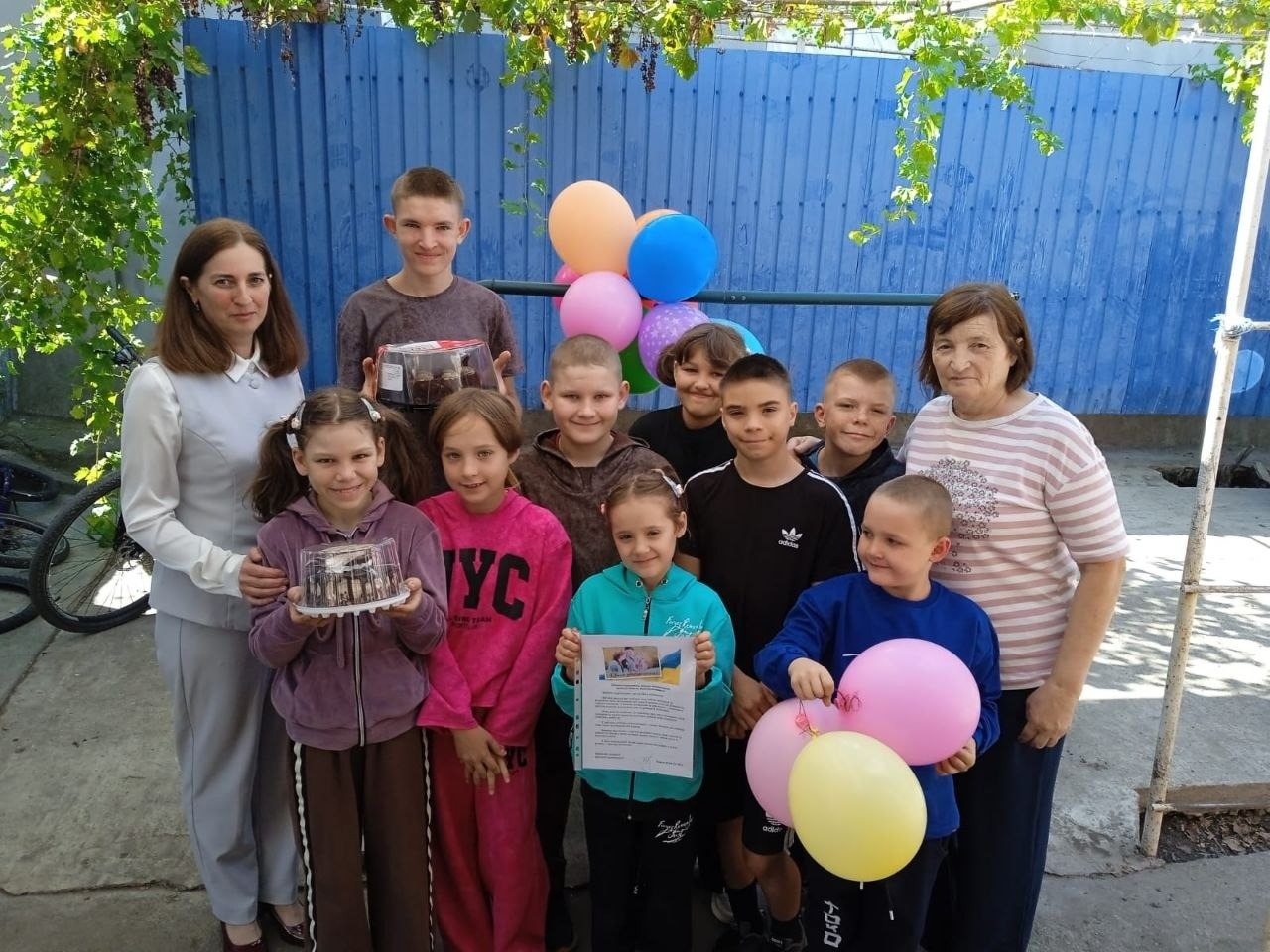Велика група дітей різного віку разом із двома дорослими жінками позує для спільного фото на подвір'ї на тлі синього паркану, прикрашеного різноколірними повітряними кульками. Ліворуч стоїть жінка у сірому костюмі, поруч із нею діти тримають у руках два солодкі торти в прозорих пластикових упаковках, дівчинка по центру демонструє офіційне привітання з прапором України, а праворуч стоїть літня жінка у смугастій футболці. Святковий візит організовано в межах соціальної підтримки, оскільки у громадах Херсонщини відбулись заходи до Дня усиновлення, під час яких фахівці служби у справах дітей Калинівської селищної ради відвідали прийомну родину та дитячий будинок сімейного типу, щоб поспілкуватися та пригостити вихованців тортами.