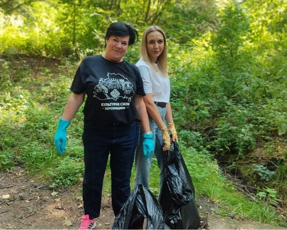 Дві жінки з числа переселенців Херсонщини та фахівців координаційного центру «Вільні разом» у Чернівцях стоять на лісовій стежці парку «Жовтневий» під час всеукраїнської толоки «World Cleanup Day 2026». Ліворуч стоїть жінка у чорній футболці з білим принтом «КУЛЬТУРНІ СИЛИ ХЕРСОНЩИНИ», темних штанях та яскравих бірюзових рукавичках, а праворуч — дівчина у білій футболці та блакитних джинсах, яка тримає великий чорний поліетиленовий пакет для збору сміття на тлі густих зелених дерев і кущів паркової зони, де також долучилися представники Палацу молоді і студентів Херсонської обласної ради.