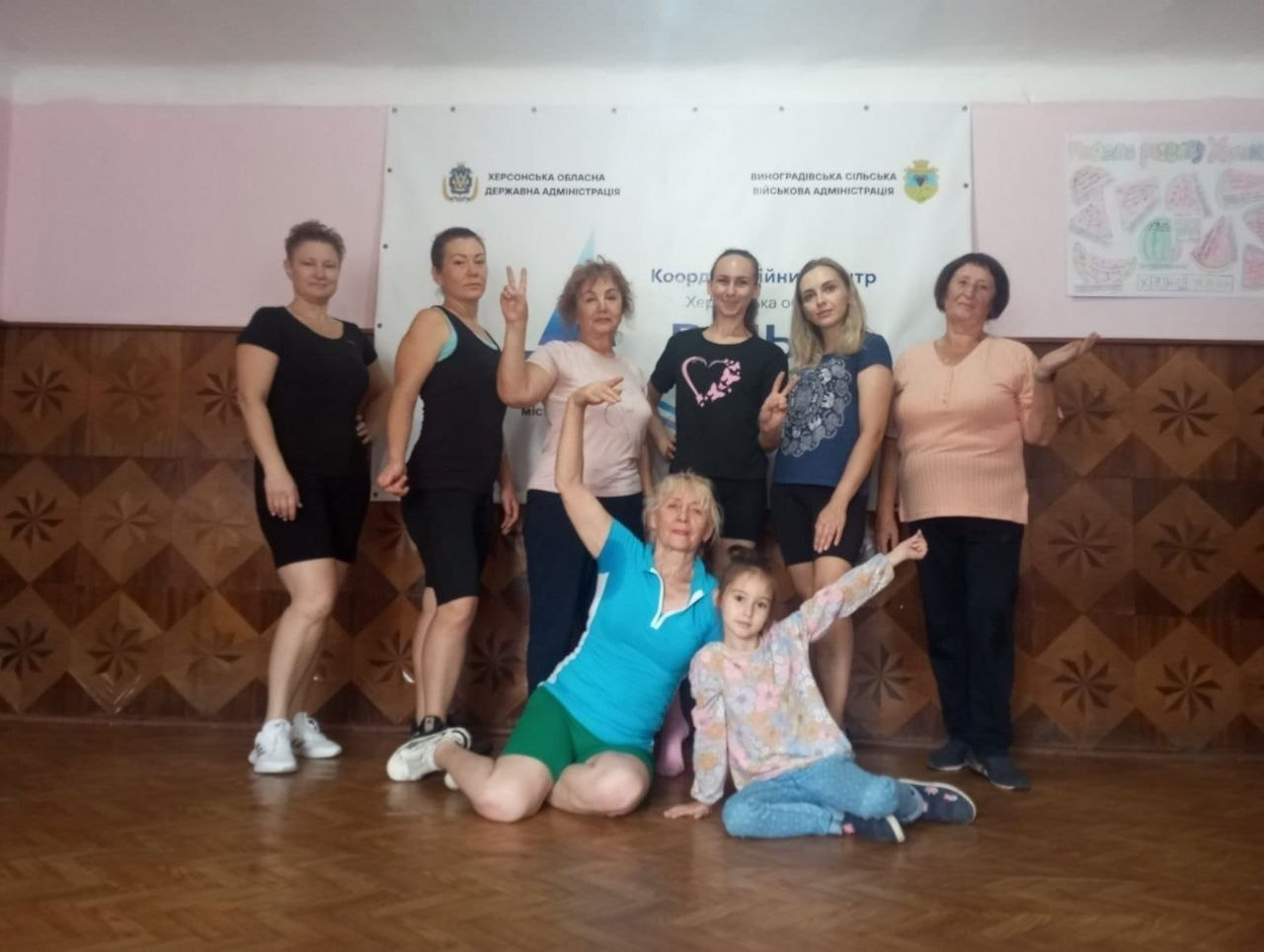 Сім жінок та одна дівчинка з числа переселенців Херсонщини позують для спільного фото в залі на тлі великого офіційного банера після спортивного тренування «Будь сильною», яке провела фітнес-тренерка Ірина Котова. Настільний білий банер містить написи «ХЕРСОНСЬКА ОБЛАСНА ДЕРЖАВНА АДМІНІСТРАЦІЯ», «ВИНОГРАДІВСЬКА СІЛЬСЬКА ВІЙСЬКОВА АДМІНІСТРАЦІЯ» та логотип координаційного центру «Вільні разом» у Чернівцях. Учасниці заходу одягнені у спортивні футболки, шорти та лосини: п'ять жінок стоять у ряд, показуючи жест перемоги, одна жінка та дитина позитивно позують сидячи на дерев'яному паркеті попереду, а ще одна учасниця стоїть ліворуч на тлі стіни з коричневими дерев'яними панелями.
