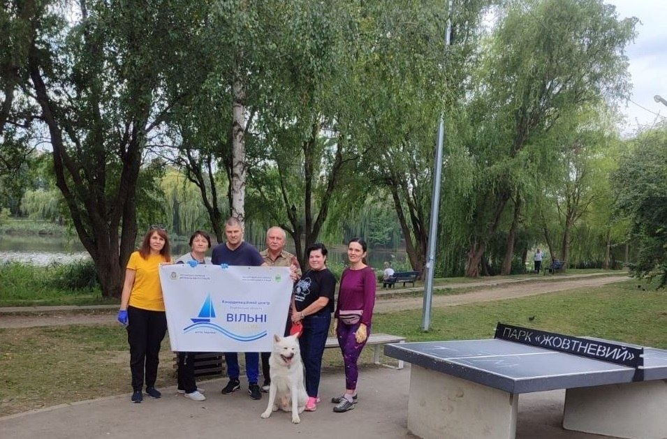 Група переселенців із Херсонщини позує для спільного фото в парку «Жовтневий» у Чернівцях під час квесту «Моє місто — Херсон» до Дня Херсона, проведеного координаційним центром «Вільні разом» спільно з Палацом молоді і студентів Херсонської обласної ради. Шестеро дорослих учасників у зручному повсякденному одязі та захисних рукавичках вишикувалися в ряд на асфальтованій доріжці біля озера і тримають великий білий банер із синім вітрильником та написом «Координаційний центр підтримки цивільного населення Херсонської області ВІЛЬНІ РАЗОМ», а попереду них сидить великий білий собака. Праворуч на передньому плані розташований бетонний тенісний стіл із написом «ПАРК «ЖОВТНЕВИЙ» на сітці на тлі зелених дерев і паркової зони.