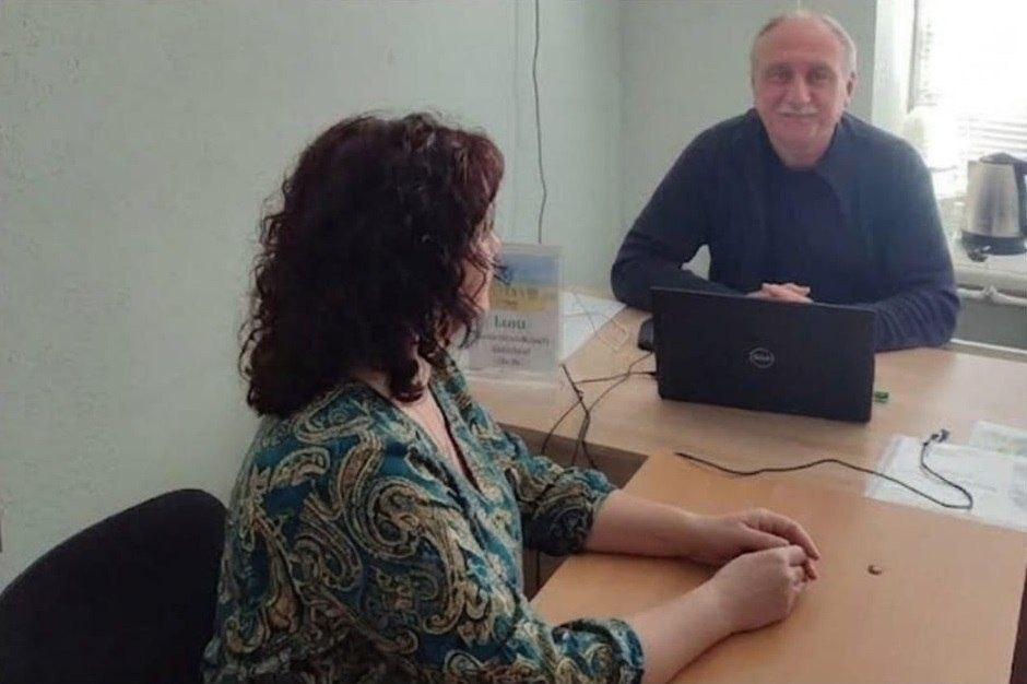 Юрист-волонтер консультує жінку з числа переселенців із Херсонщини в кабінеті координаційного центру «Вільні разом» у Чернівцях під час надання юридичної допомоги. На задньому плані обличчям до камери сидить усміхнений сивочолий чоловік у темно-синьому лонгсліві, перед яким на столі відкритий чорний ноутбук Dell, а на передньому плані спиною та боком до кадру розмістилася жінка з пишним кучерявим темним волоссям у синьо-зеленій блузі з візерунком «пейслі», яка склала руки на столі й уважно слухає фахівця на тлі настільної таблички хабу.