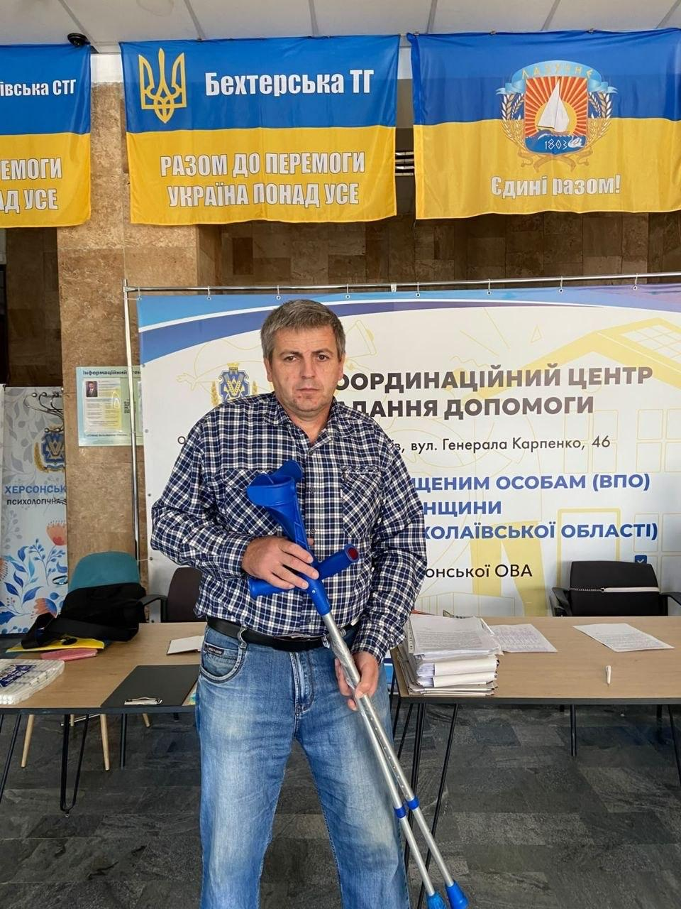 Чоловік середнього віку у синьо-білій картатій сорочці та блакитних джинсах тримає в руках нові металеві милиці з синіми пластиковими елементами, які він отримав як допоміжні засоби реабілітації в миколаївському координаційному центрі «Вільні разом» від організації «Фонд громади міста Херсон «Захист». Переселенець із Херсонщини стоїть у приміщенні хабу на тлі великого інформаційного банера «КООРДИНАЦІЙНИЙ ЦЕНТР НАДАННЯ ДОПОМОГИ ПЕРЕМІЩЕНИМ ОСОБАМ (ВПО) ХЕРСОНЩИНИ В МИКОЛАЇВСЬКІЙ ОБЛАСТІ». Під стелею над ним закріплені синьо-жовті прапори громад, зокрема Бехтерської ТГ з написом «Разом до перемоги. Україна понад усе» та прапор із гаслом «Єдині разом!» і символікою вітрильника на тлі робочих столів із паперами.